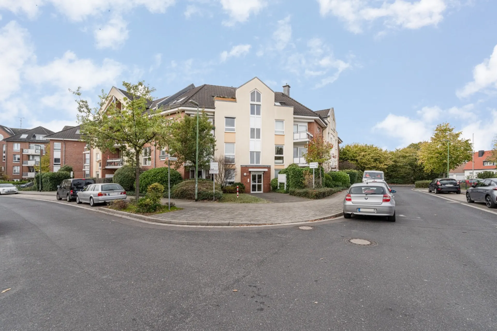 Mehrfamilienhaus an einer Straßenecke mit geparkten Autos und Bäumen im Vordergrund.