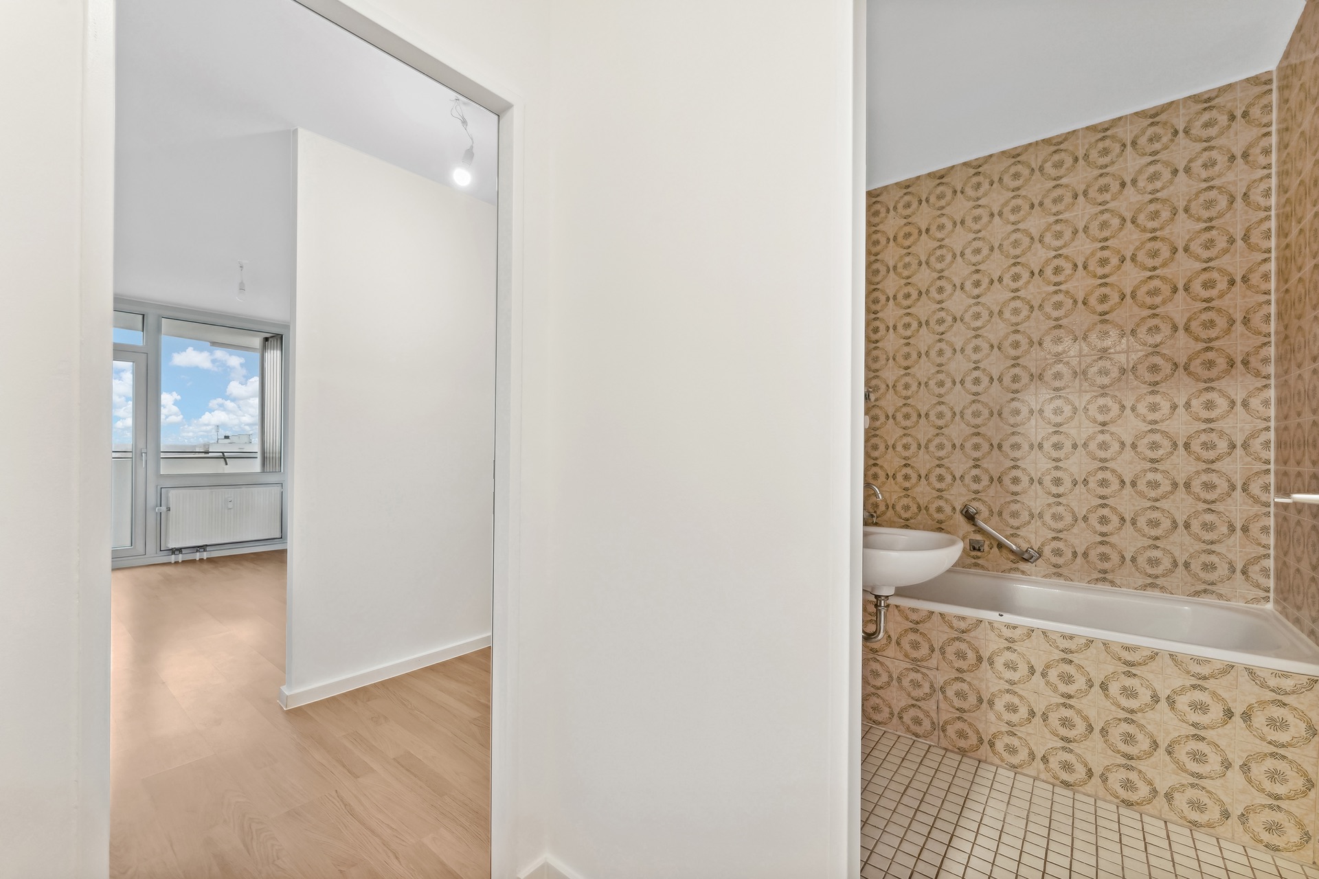 Badezimmer mit gemusterten Fliesen, Badewanne und Waschbecken neben einem hellen Raum mit Fenster.