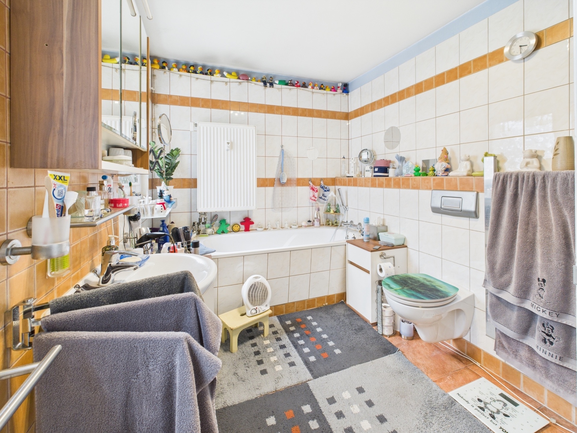 Helles Badezimmer mit Badewanne, Waschbecken, Toilette, Regalen und vielen Pflegeprodukten.