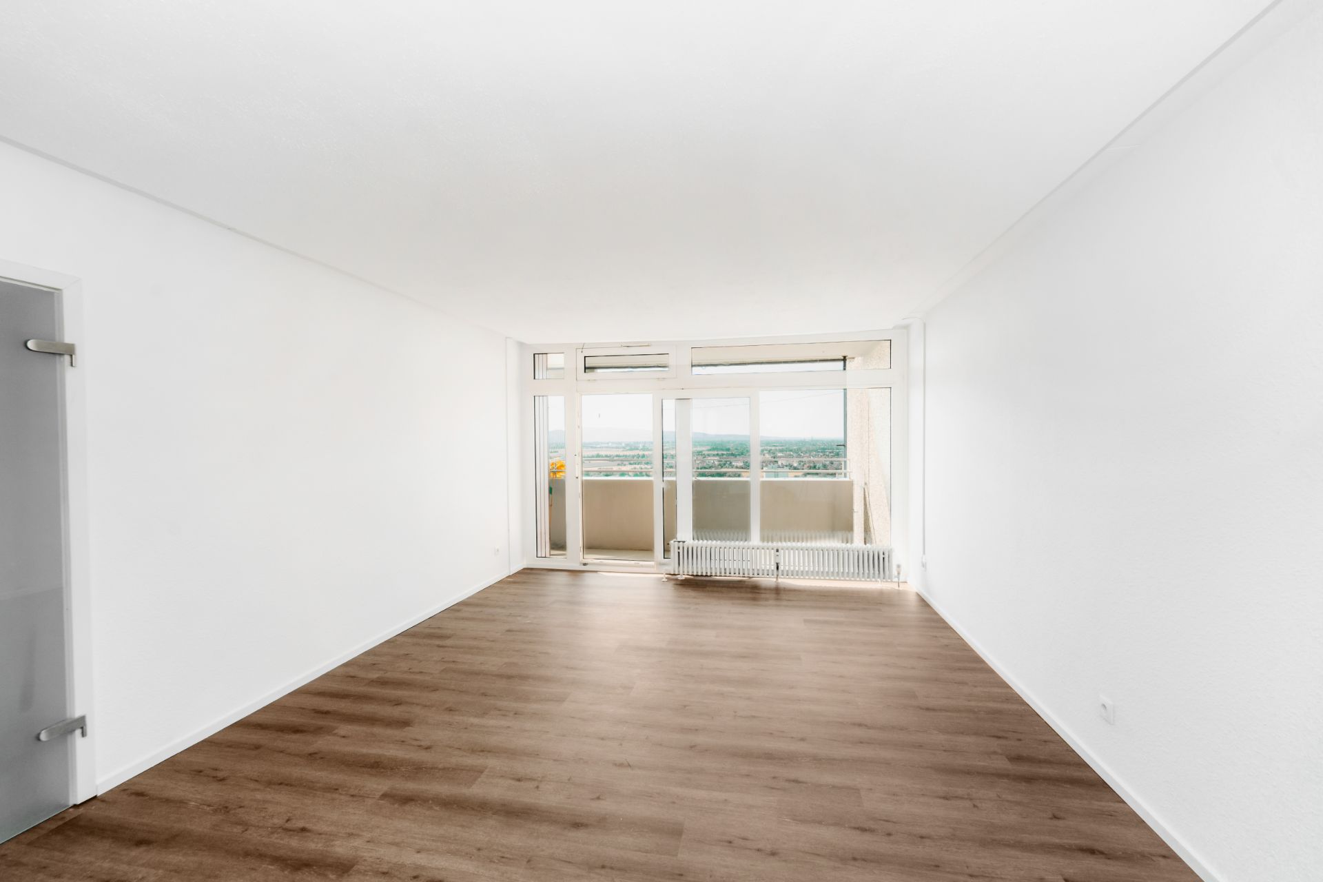 Leeres Wohnzimmer mit Holzfußboden, weißen Wänden und großer Fensterfront mit Blick nach draußen.