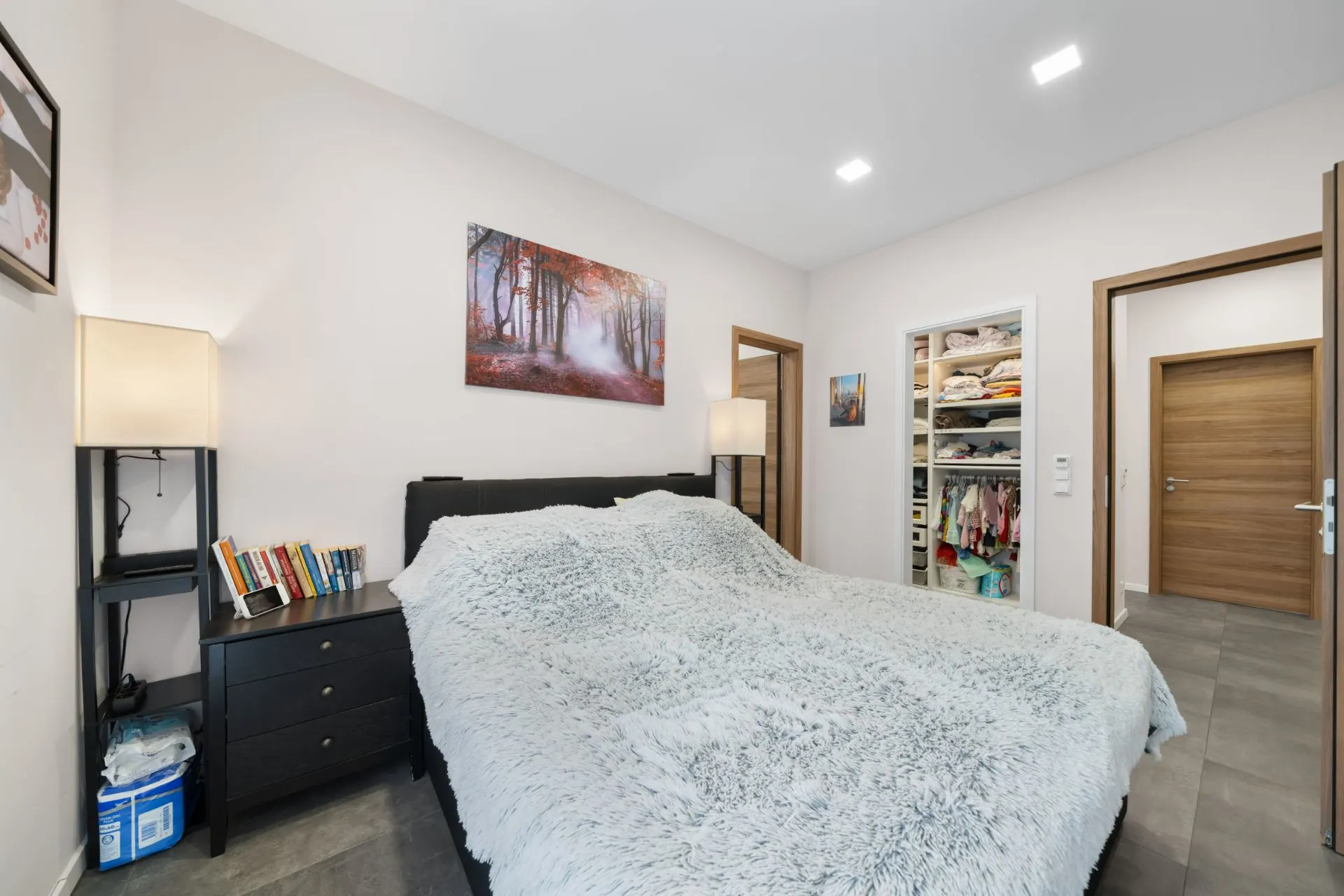 Schlafzimmer mit großem Bett, Nachttisch, Regal, offenem Kleiderschrank und Wandbild.