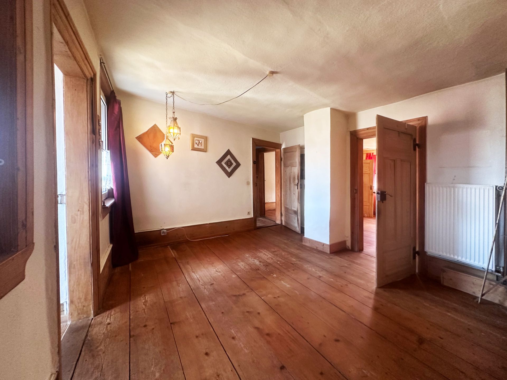 Helles Zimmer mit Holzboden, offenen Türen, Wanddekoration und Fenstern mit Vorhängen.