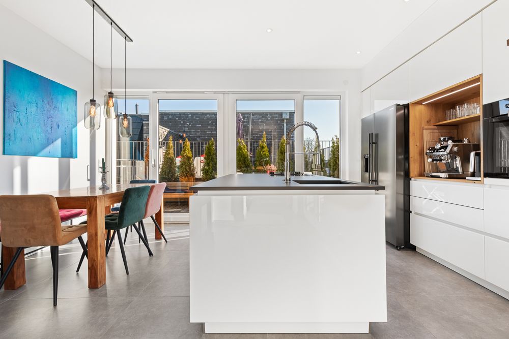 Moderne Küche mit Kochinsel, Esstisch und bunten Stühlen, große Fenster im Hintergrund.