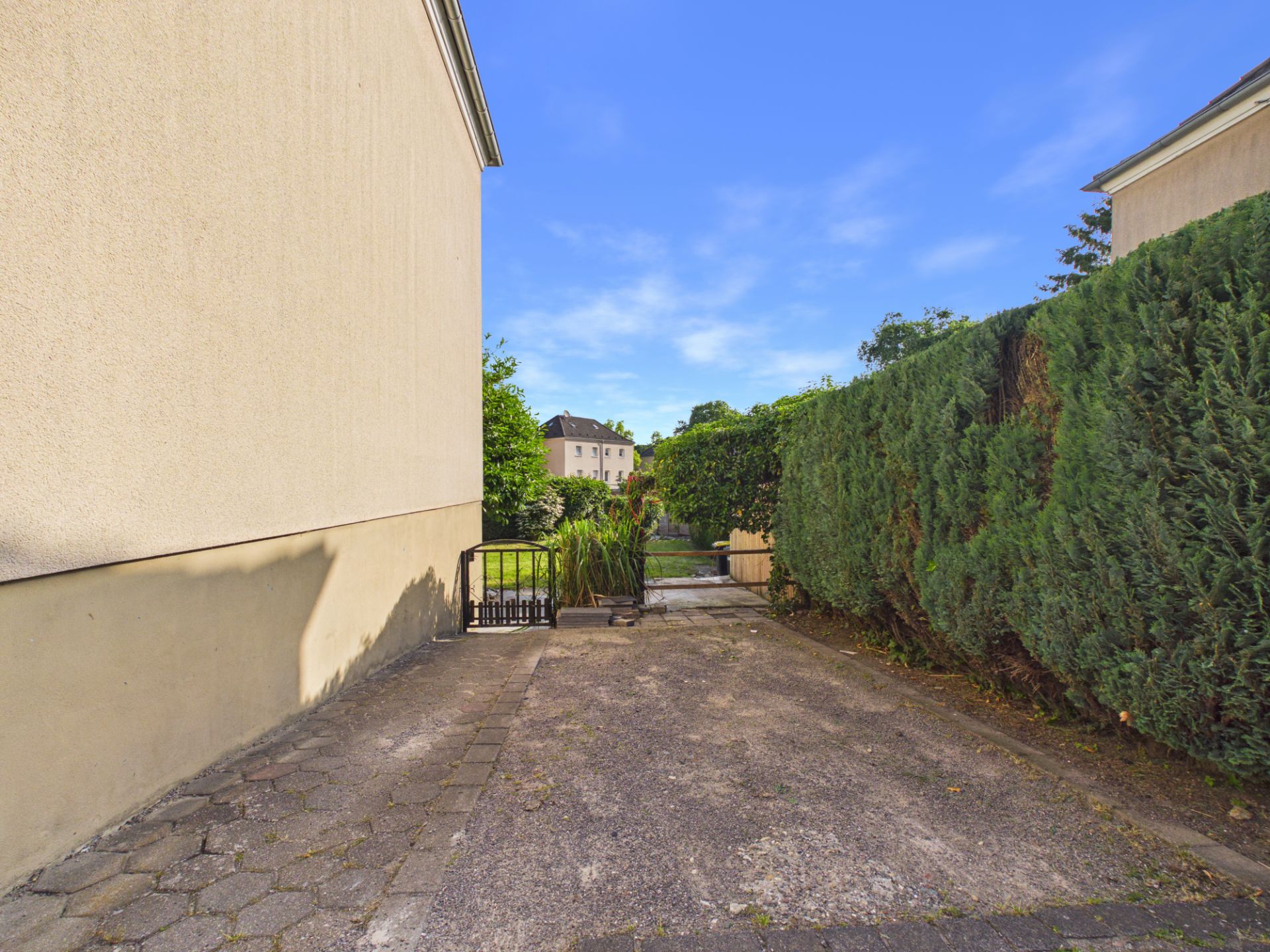 Schmaler gepflasterter Weg zwischen Hauswand und hoher Hecke mit kleinem Tor am Ende.