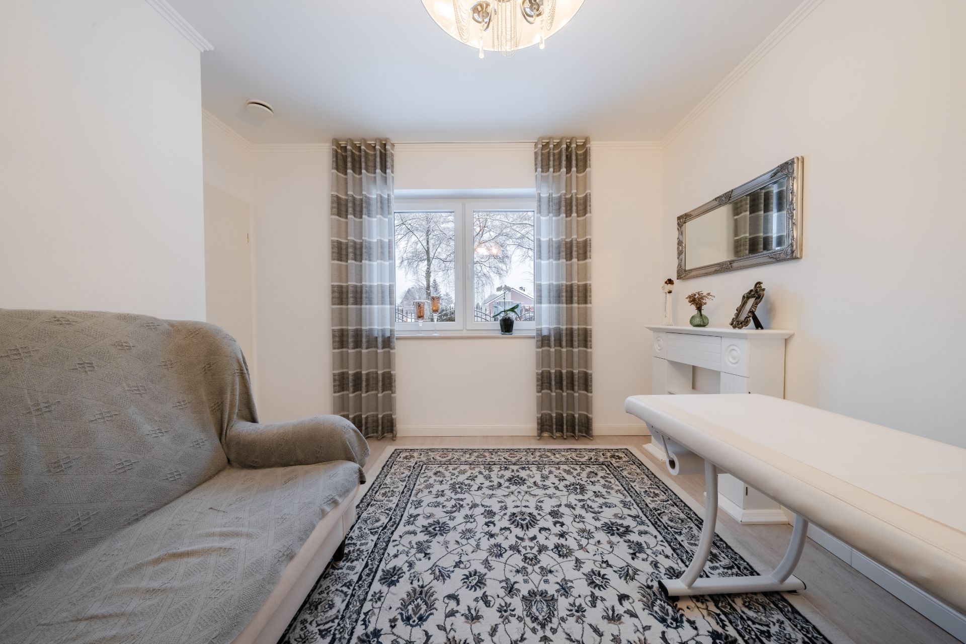 Helles Zimmer mit Sofa, Teppich, Fenster mit Vorhängen und einem weißen Tisch an der Wand.