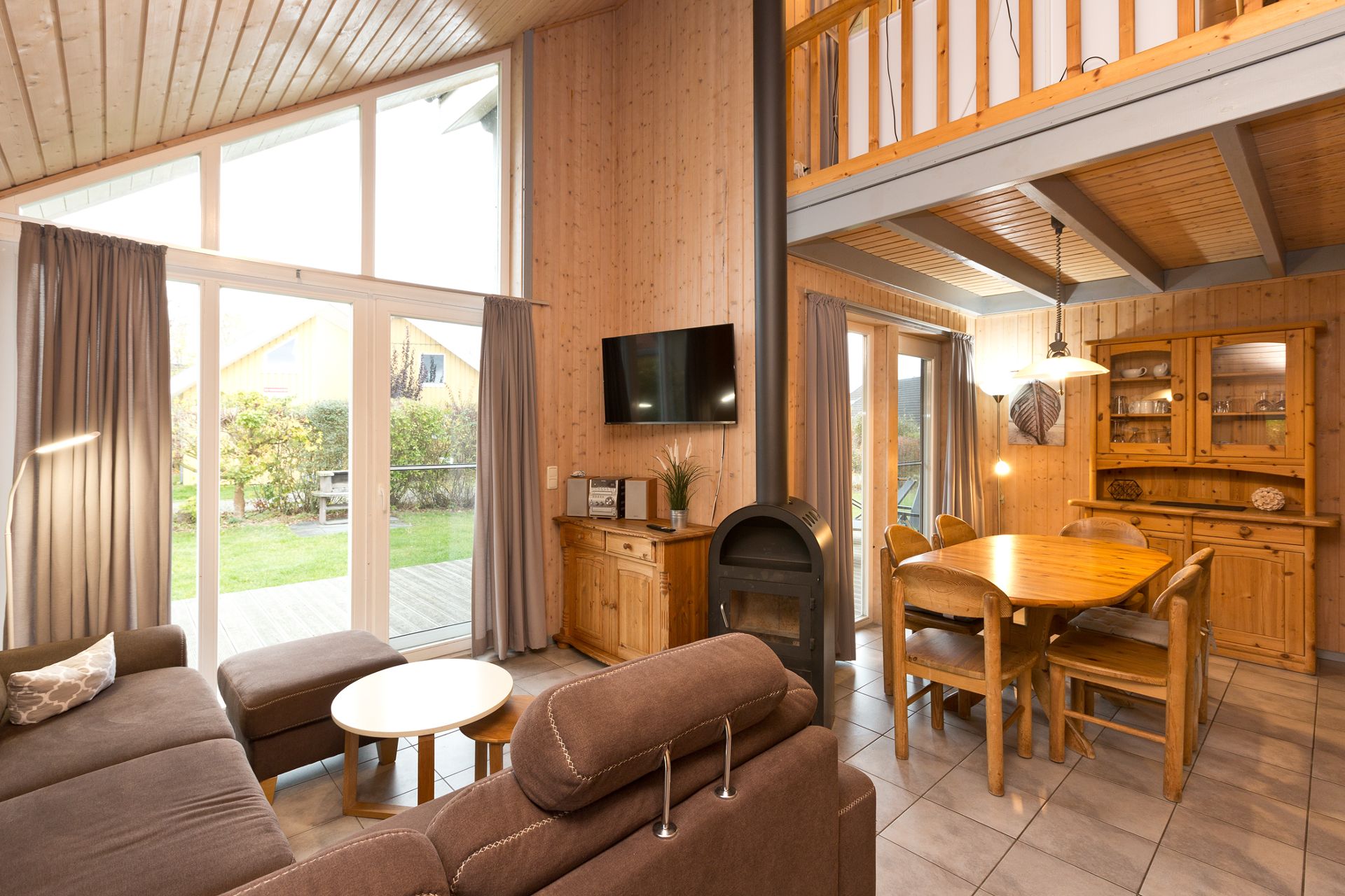 Wohnzimmer mit braunem Sofa, Esstisch, Kaminofen und großen Fenstern zum Garten.