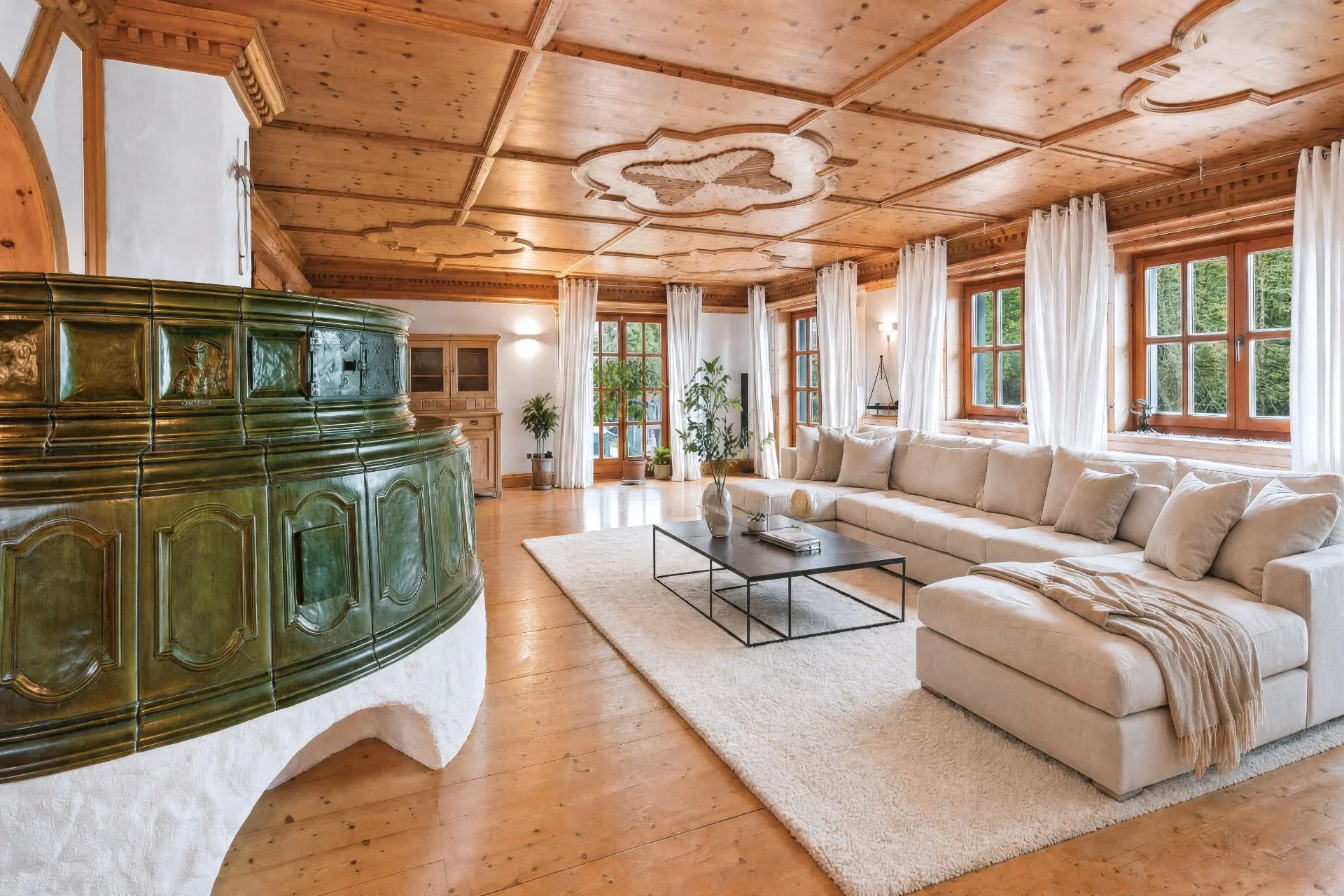 Helles Wohnzimmer mit großem Ecksofa, Kaminofen und vielen Fenstern mit weißen Vorhängen.