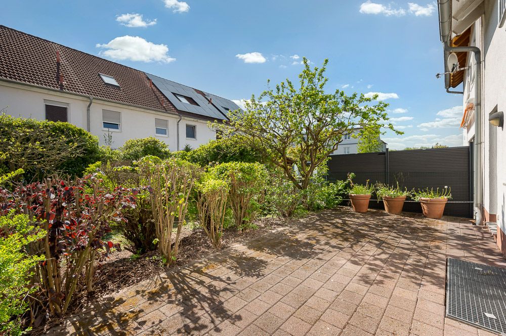 Gepflasterte Terrasse mit mehreren Pflanzen, Büschen und Baum neben Wohnhaus bei Sonnenschein.