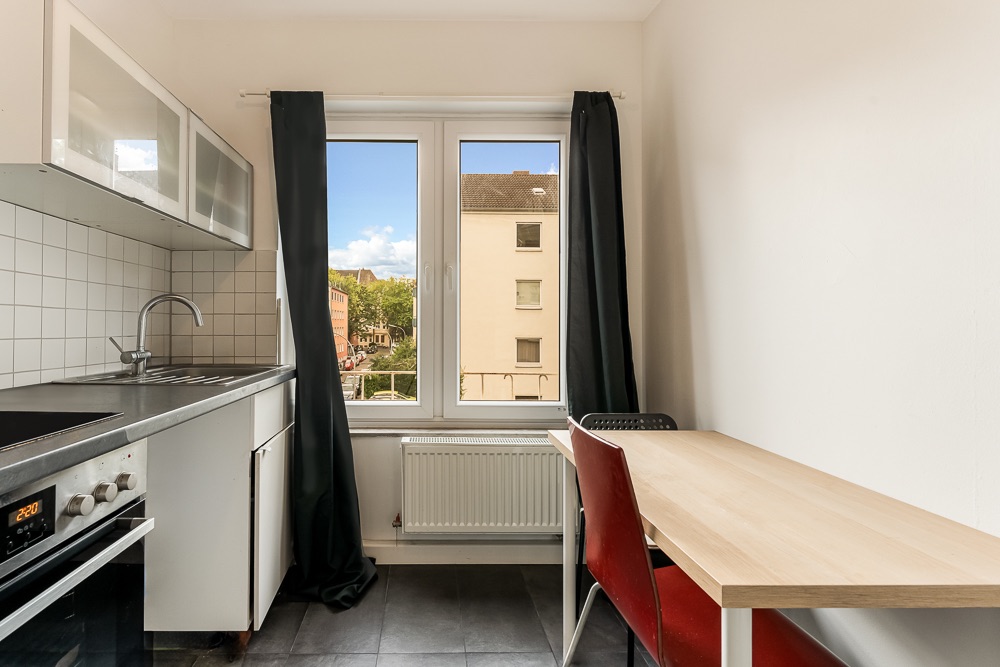 Kleine Küche mit Esstisch, zwei Stühlen, Fenster mit Vorhängen und Blick nach draußen.