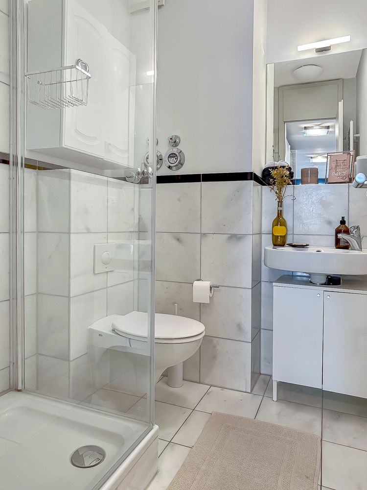 Helles Badezimmer mit Dusche, WC, Waschbecken, Spiegel und weißem Fliesendekor.