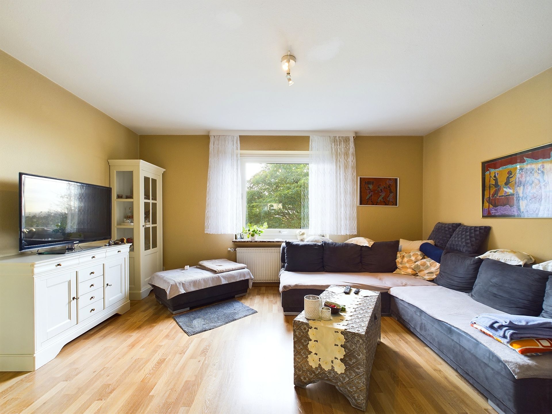 Wohnzimmer mit Ecksofa, Couchtisch, TV, Sideboard und großem Fenster mit Vorhängen.