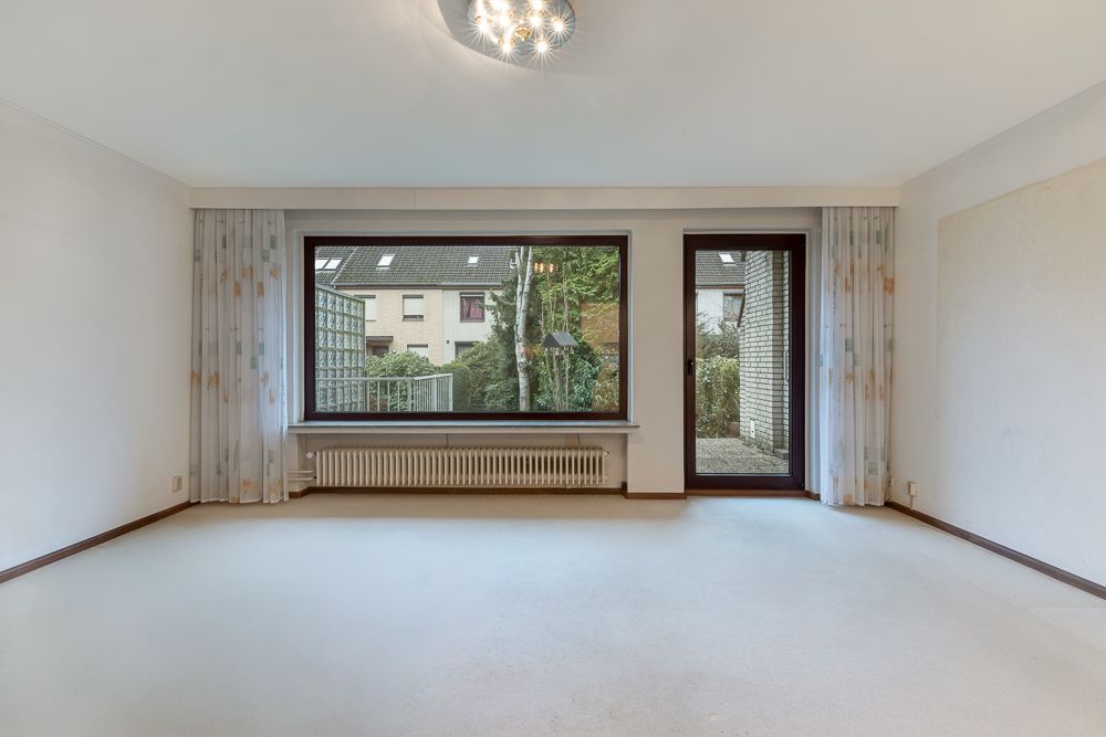 Helles, leeres Wohnzimmer mit großem Fenster, Heizung und Zugang zur Terrasse.