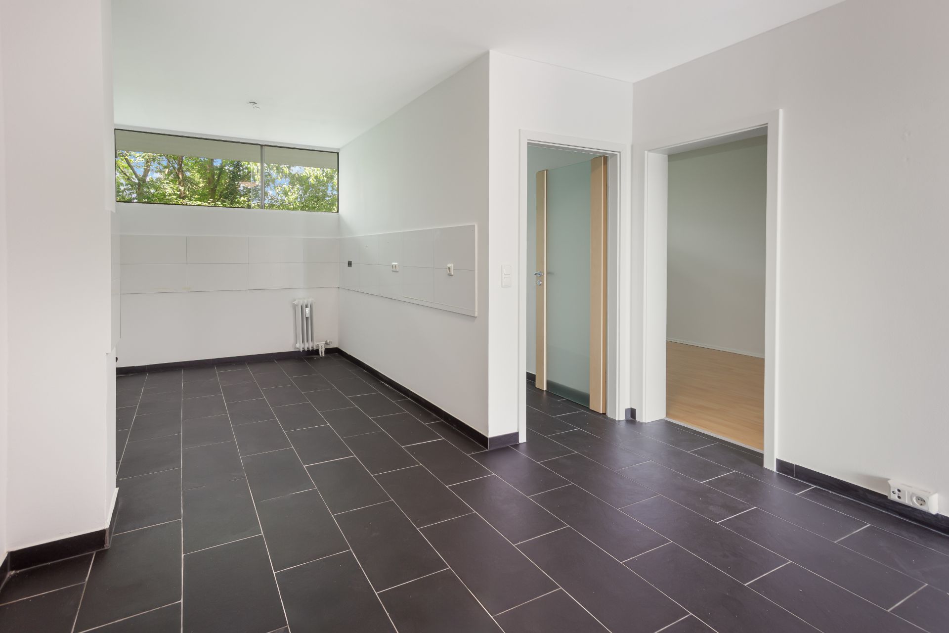 Leerer Raum mit dunklem Fliesenboden, zwei offenen Türen und Fenster mit Blick ins Grüne.