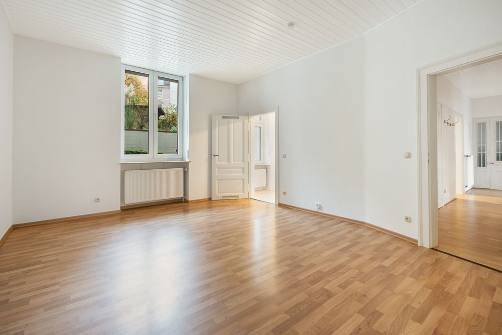 Helles leeres Zimmer mit Holzfußboden, weißen Wänden und einem großen Fenster.