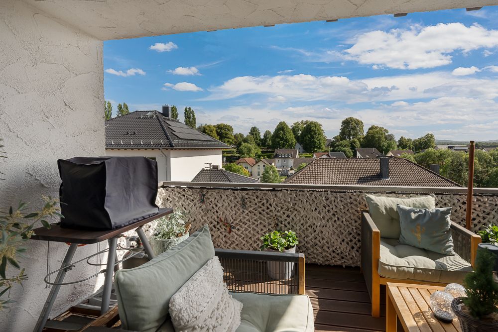 Überdachter Balkon mit Sitzmöbeln, Grill und Blick auf Dächer und Bäume.