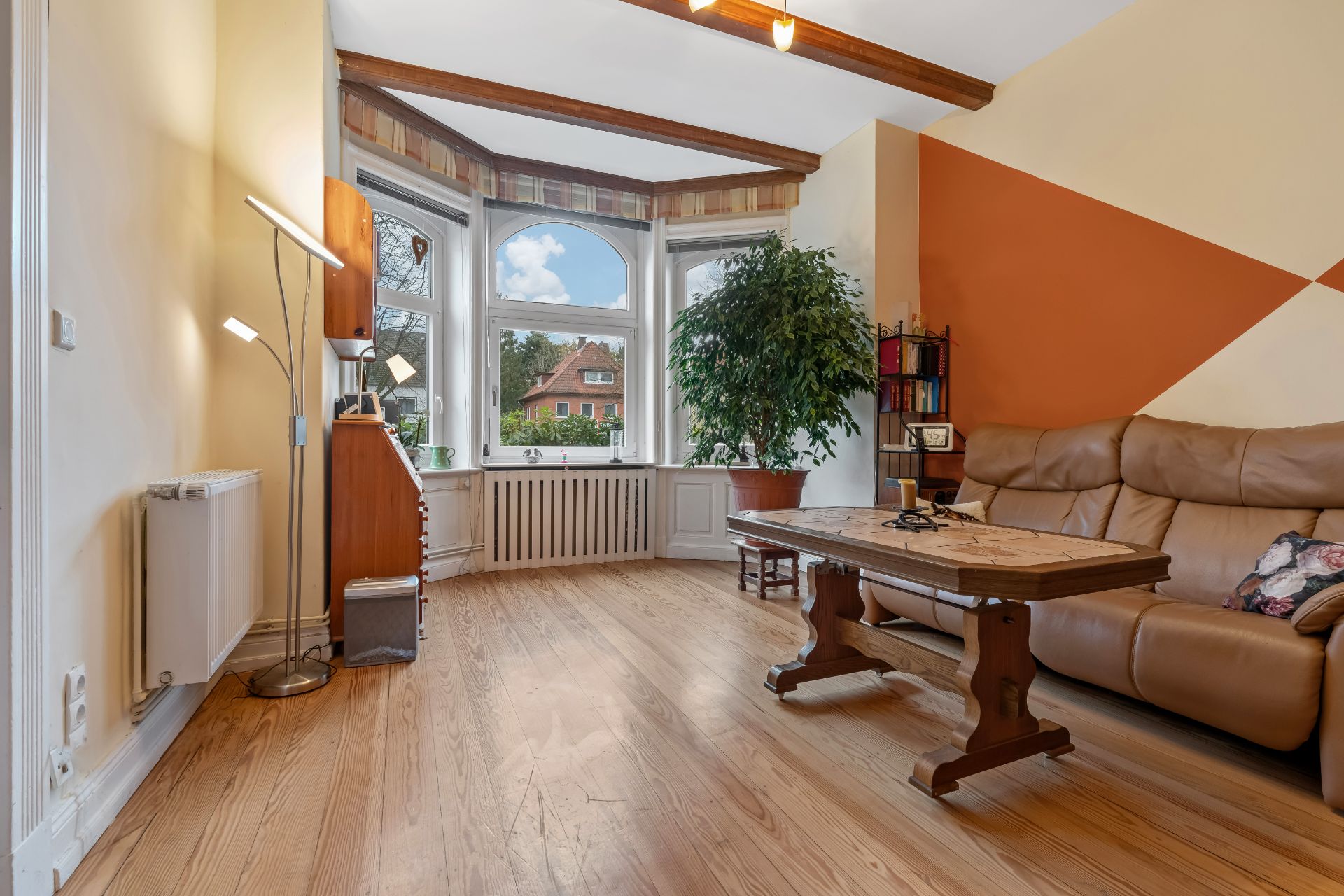 Helles Wohnzimmer mit Ledersofa, Holztisch, großer Pflanze und Erkerfenster.