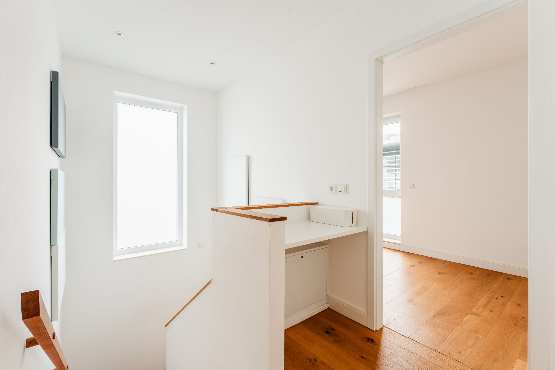 Heller Flur mit Holztreppe, großem Fenster und Durchgang zu einem Raum mit Holzboden.