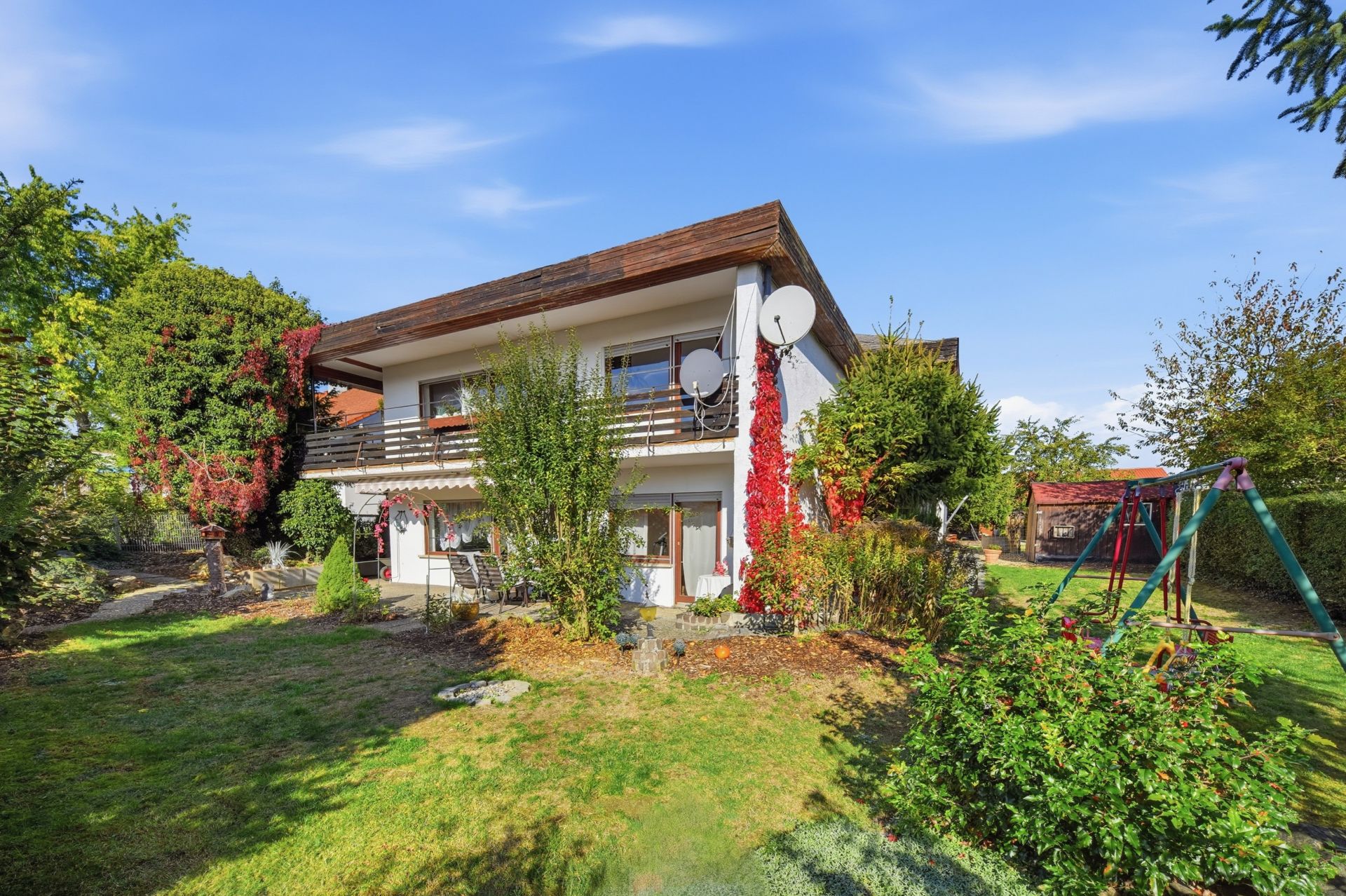 Zweistöckiges Haus mit Garten, Schaukeln und vielen Pflanzen bei sonnigem Wetter.