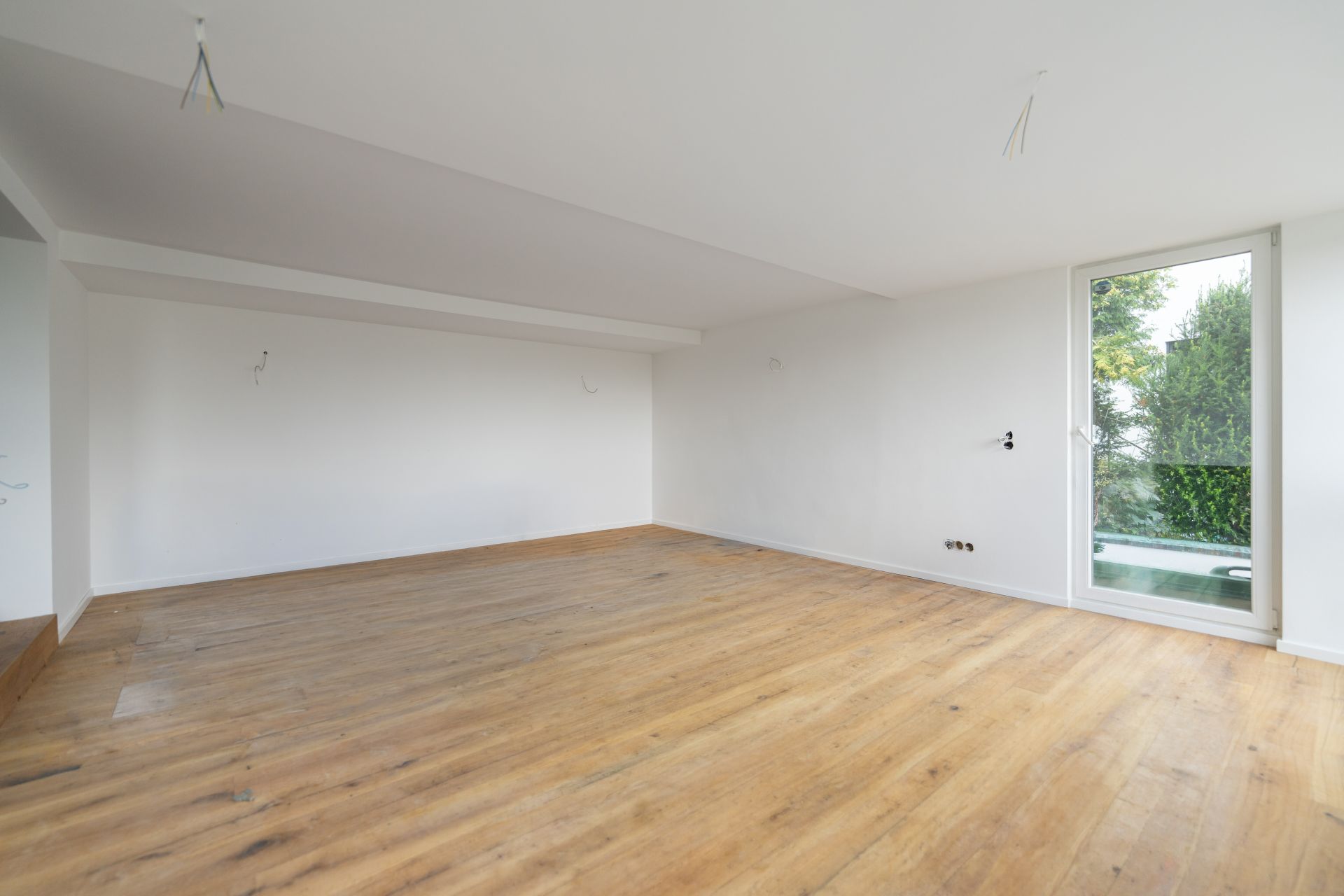 Leerer Raum mit Holzfußboden und weißer Wand, große Glastür mit Blick ins Grüne.