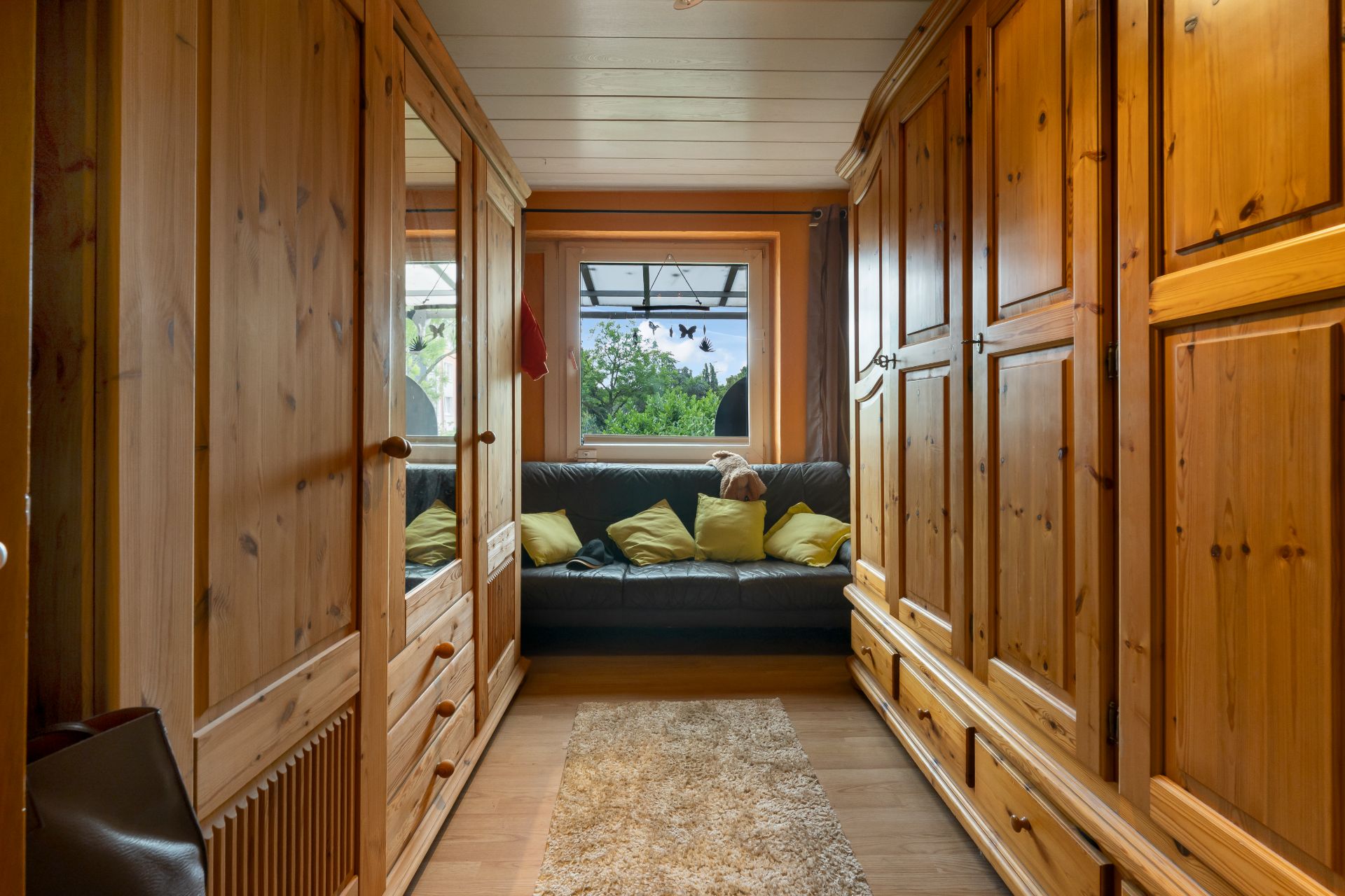 Schmaler Raum mit Holzmöbeln, Sofa am Fenster und gelben Kissen.