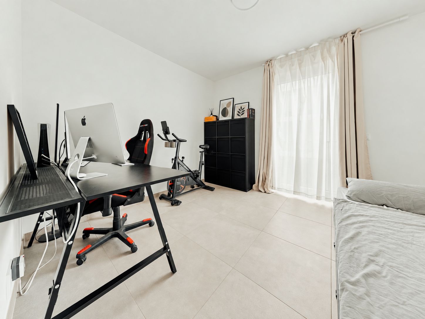 Helles Zimmer mit Schreibtisch, Computer, Stuhl, Fitnessrad, Regal und Bett.