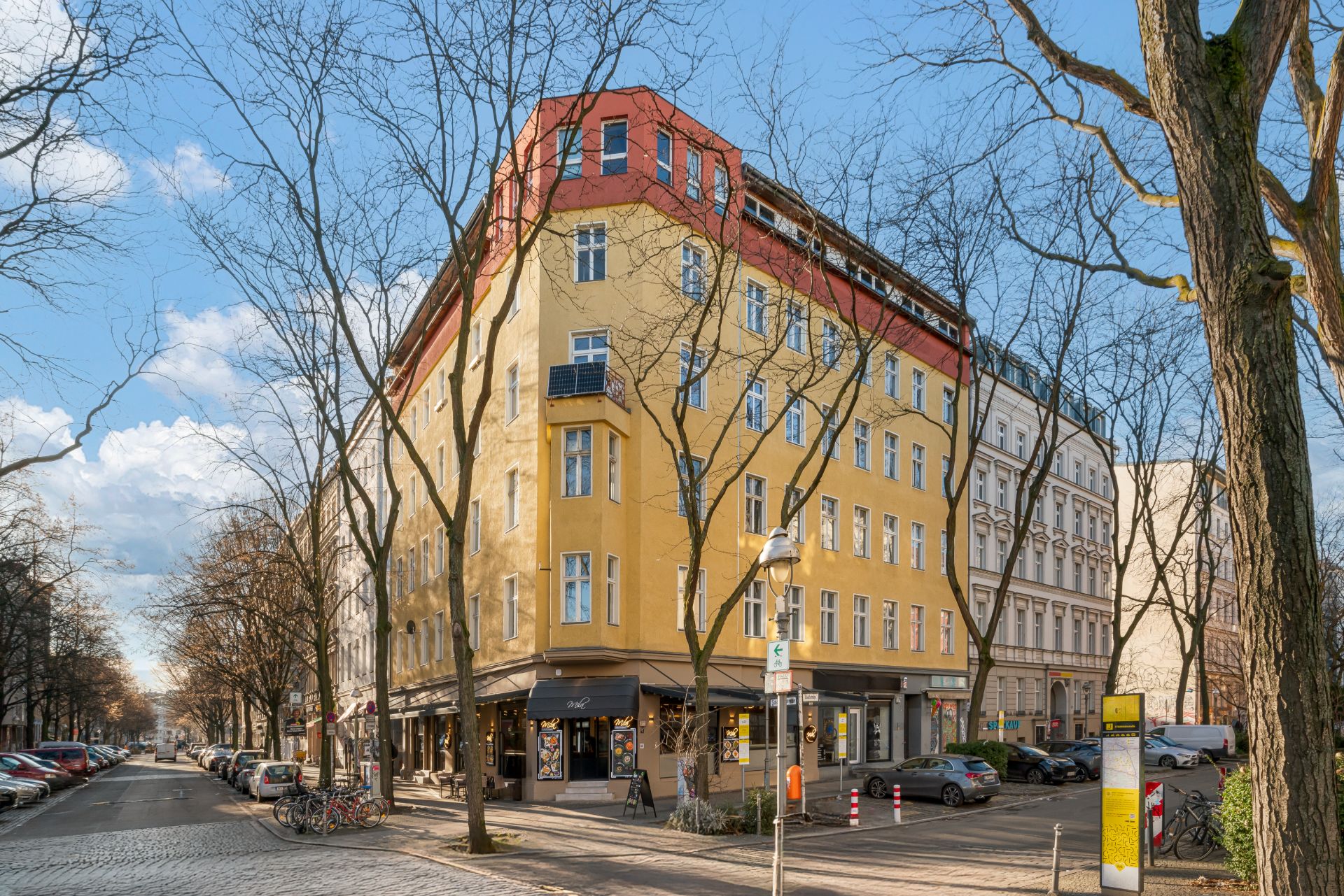 Eckhaus mit gelber Fassade und roten Akzenten, Bäume ohne Blätter, einige geparkte Autos und Fahrräder.