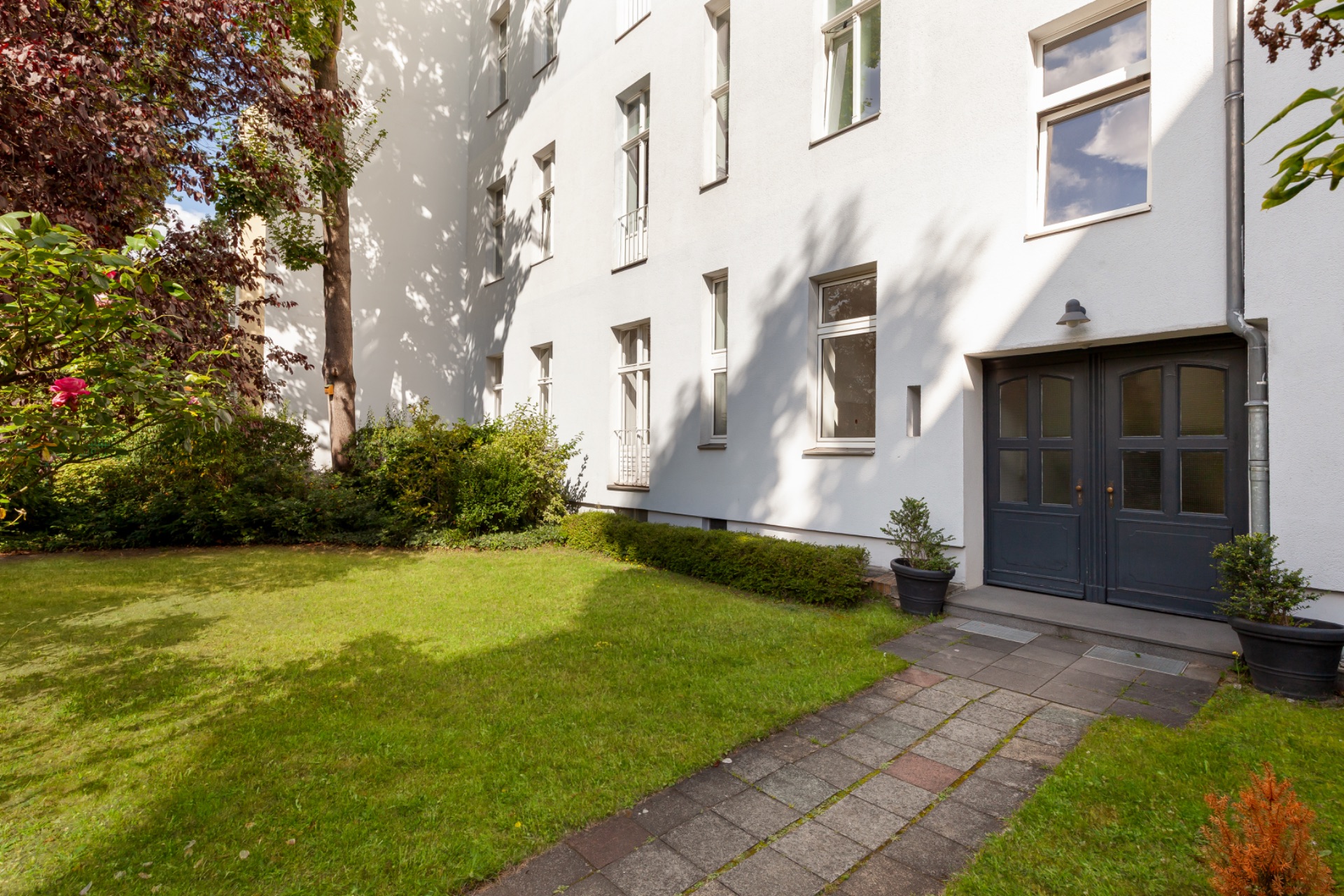 Hauseingang mit großem Fenster, Gehweg und grünem Garten mit Büschen und Bäumen.