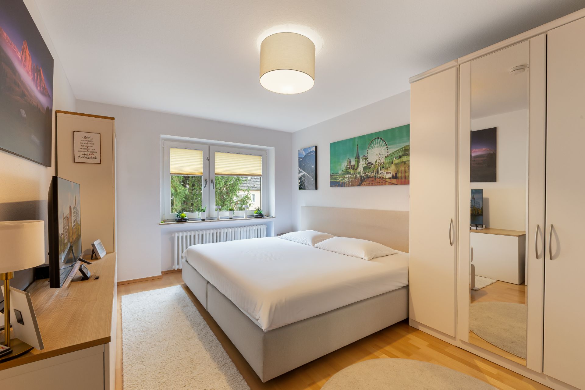 Helles Schlafzimmer mit Doppelbett, Kleiderschrank mit Spiegel und Fenstern mit Rollos.