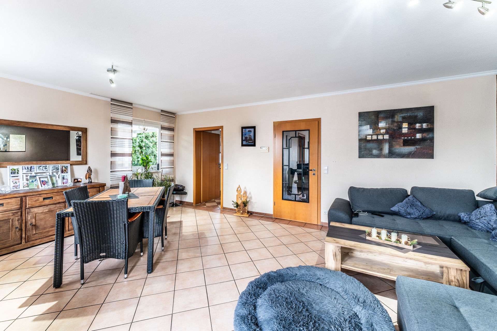 Helles Wohnzimmer mit grauem Sofa, Esstisch, Holzschrank und Dekorationen auf Fliesenboden.