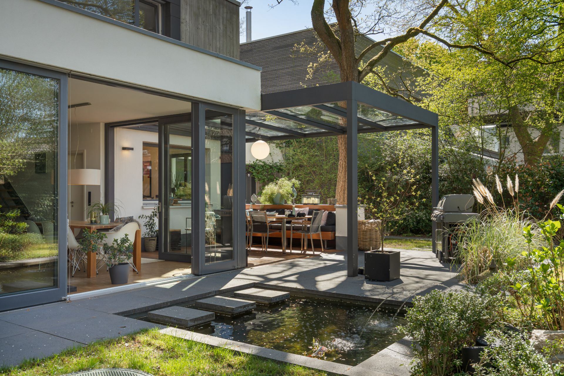 Moderne Terrasse mit Glasdach, Sitzbereich, Grill und kleinem Teich im Garten.