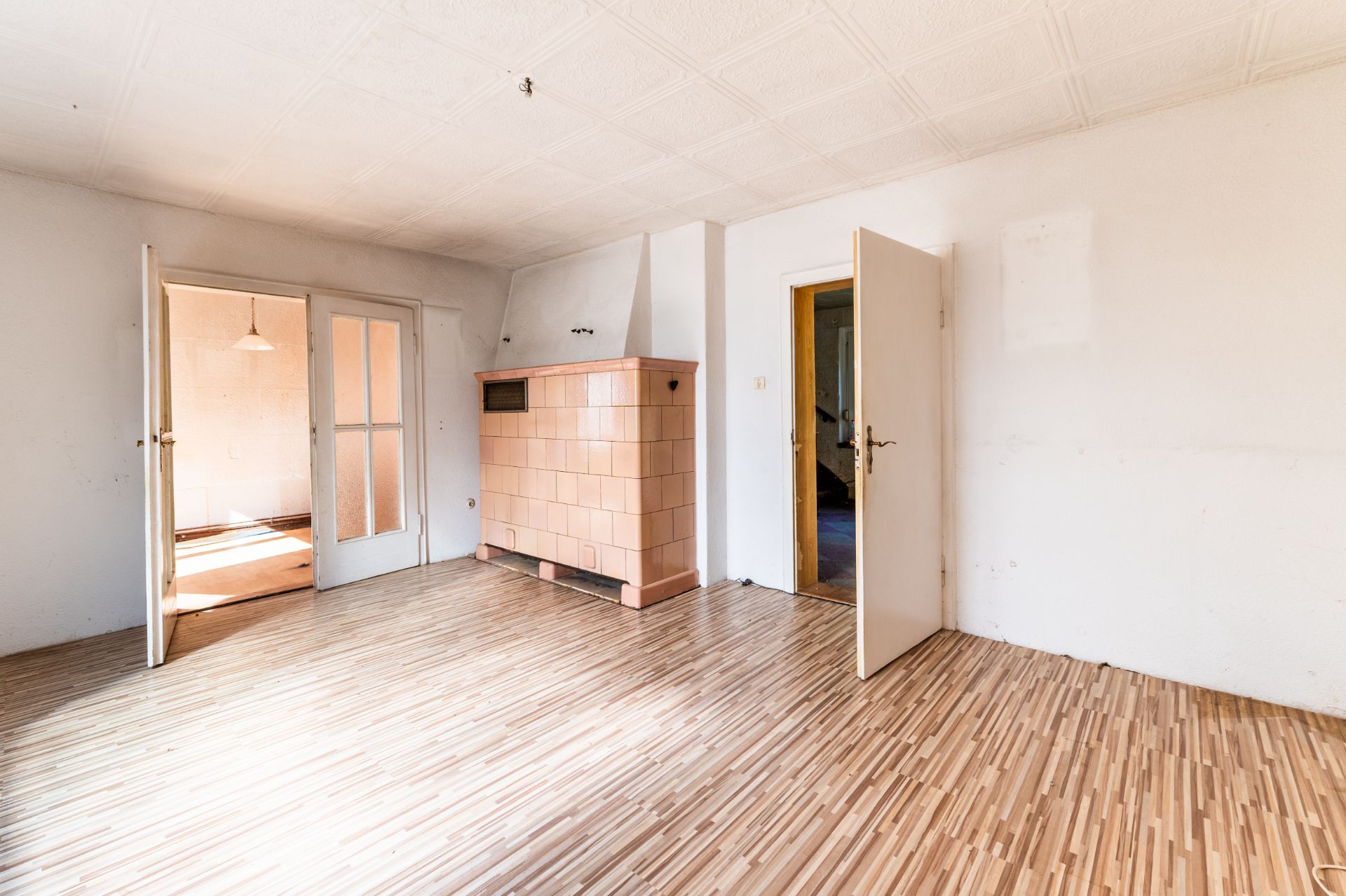 Helles Zimmer mit Holzboden, zwei geöffneten Türen und einem großen gemauerten Ofen an der Wand.