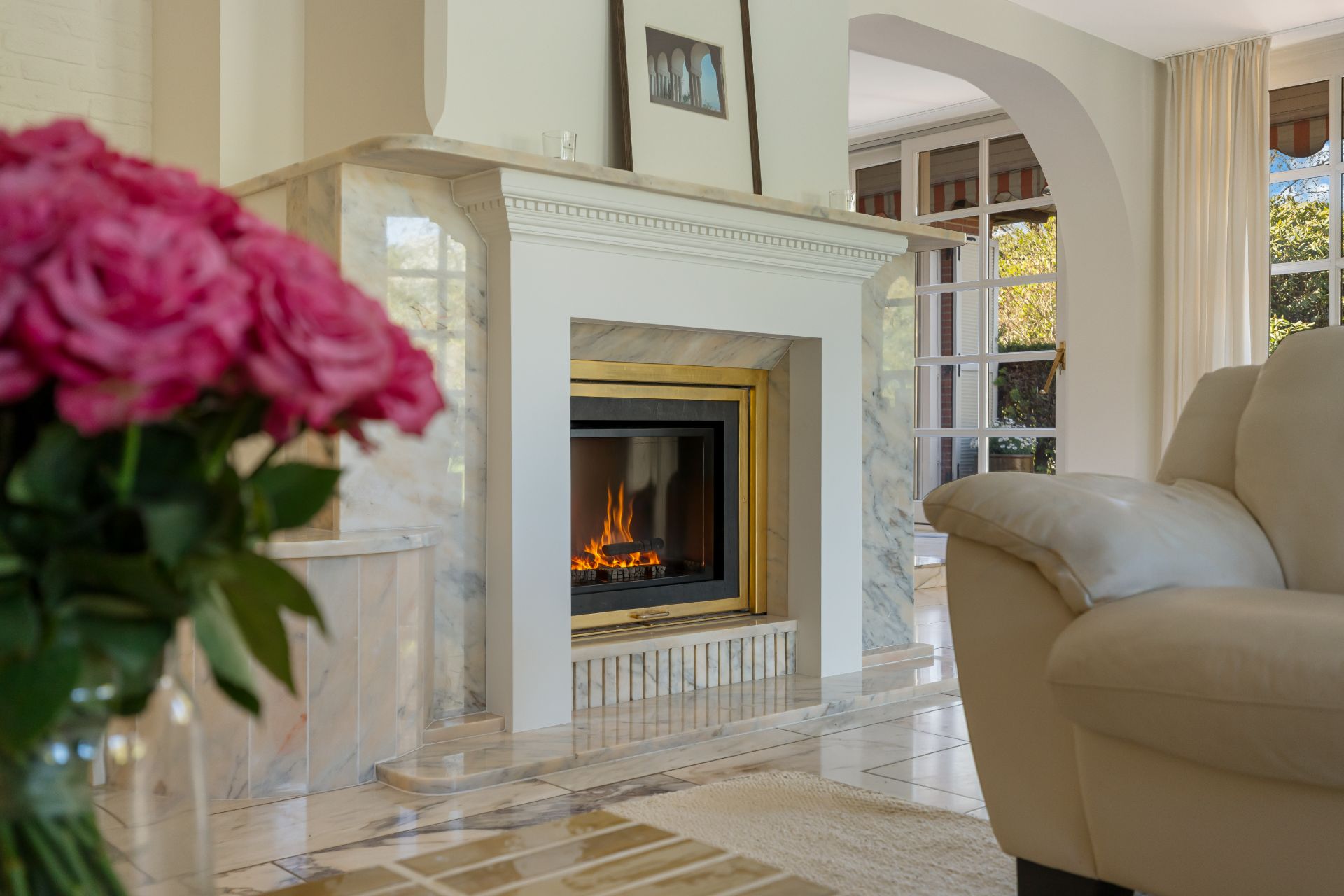 Wohnzimmer mit Kamin, beigem Sofa, Blumenstrauß im Vordergrund und großen Fenstern im Hintergrund.