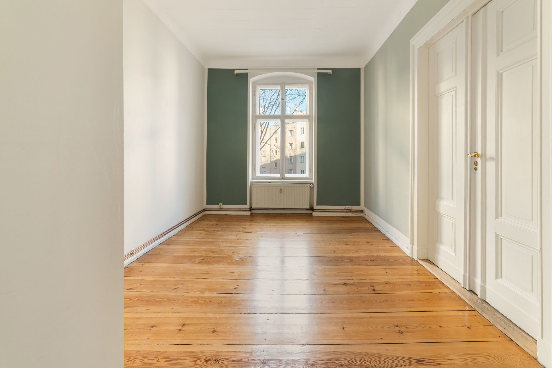 Helles, leeres Zimmer mit Holzboden, weißer Tür und großem Fenster.