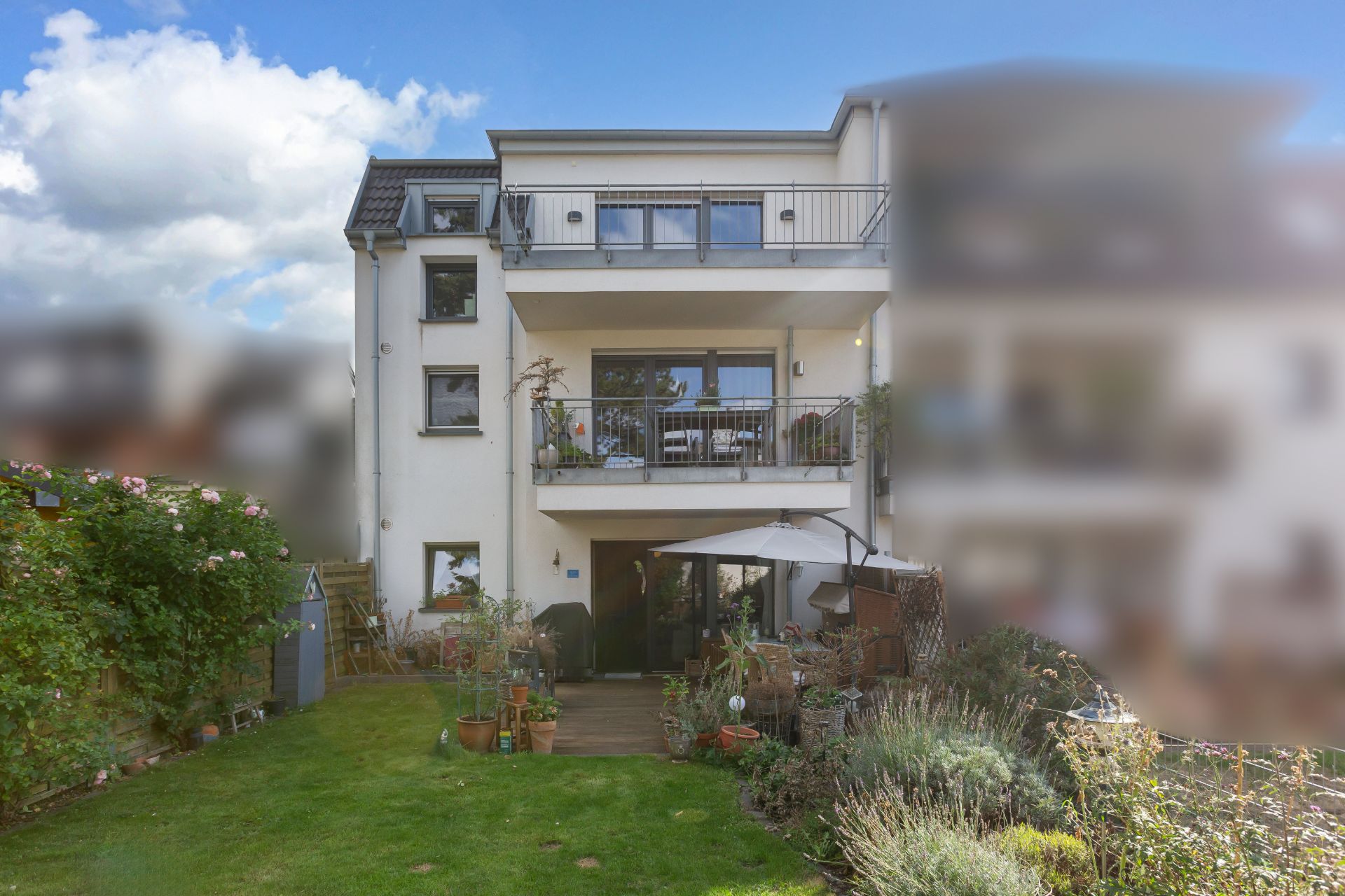 Mehrstöckiges Wohnhaus mit Balkon, Terrasse, Garten und Sitzbereich unter einem Sonnenschirm.