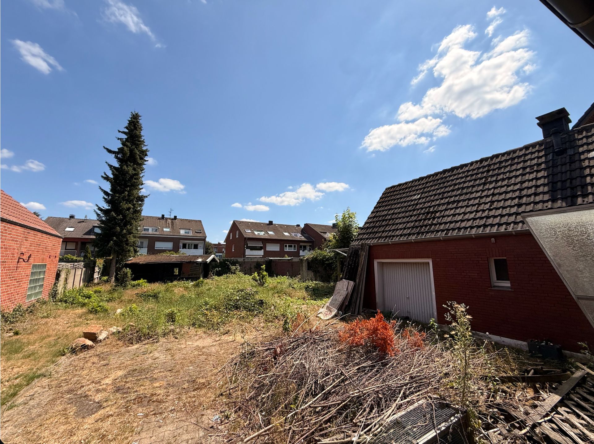Garten mit etwas Unkraut, Sträuchern und einem roten Nebengebäude mit Garage an einem sonnigen Tag.