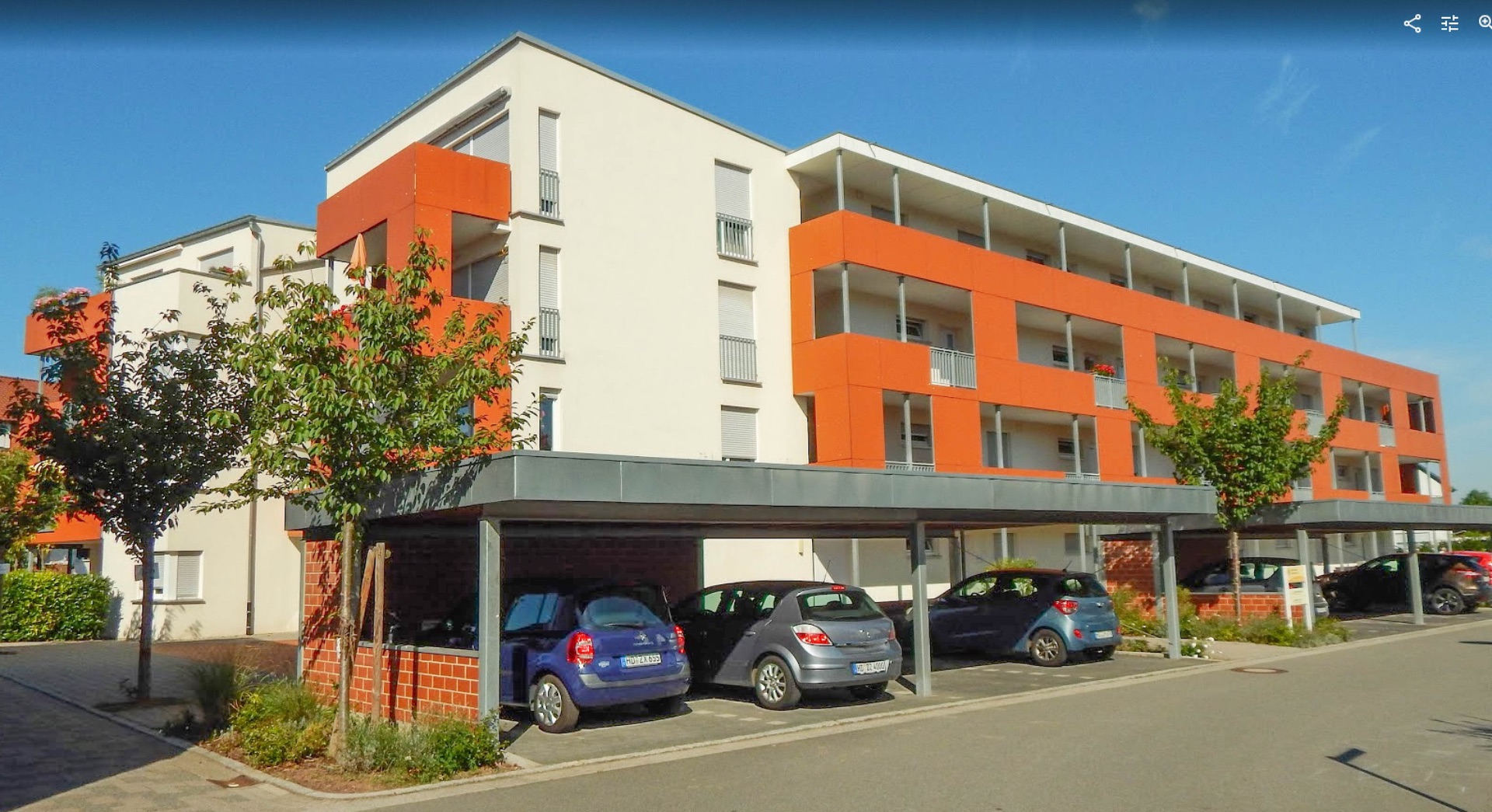 Modernes Wohngebäude mit Balkonen und Parkplätzen unter Carports, einige Bäume davor.