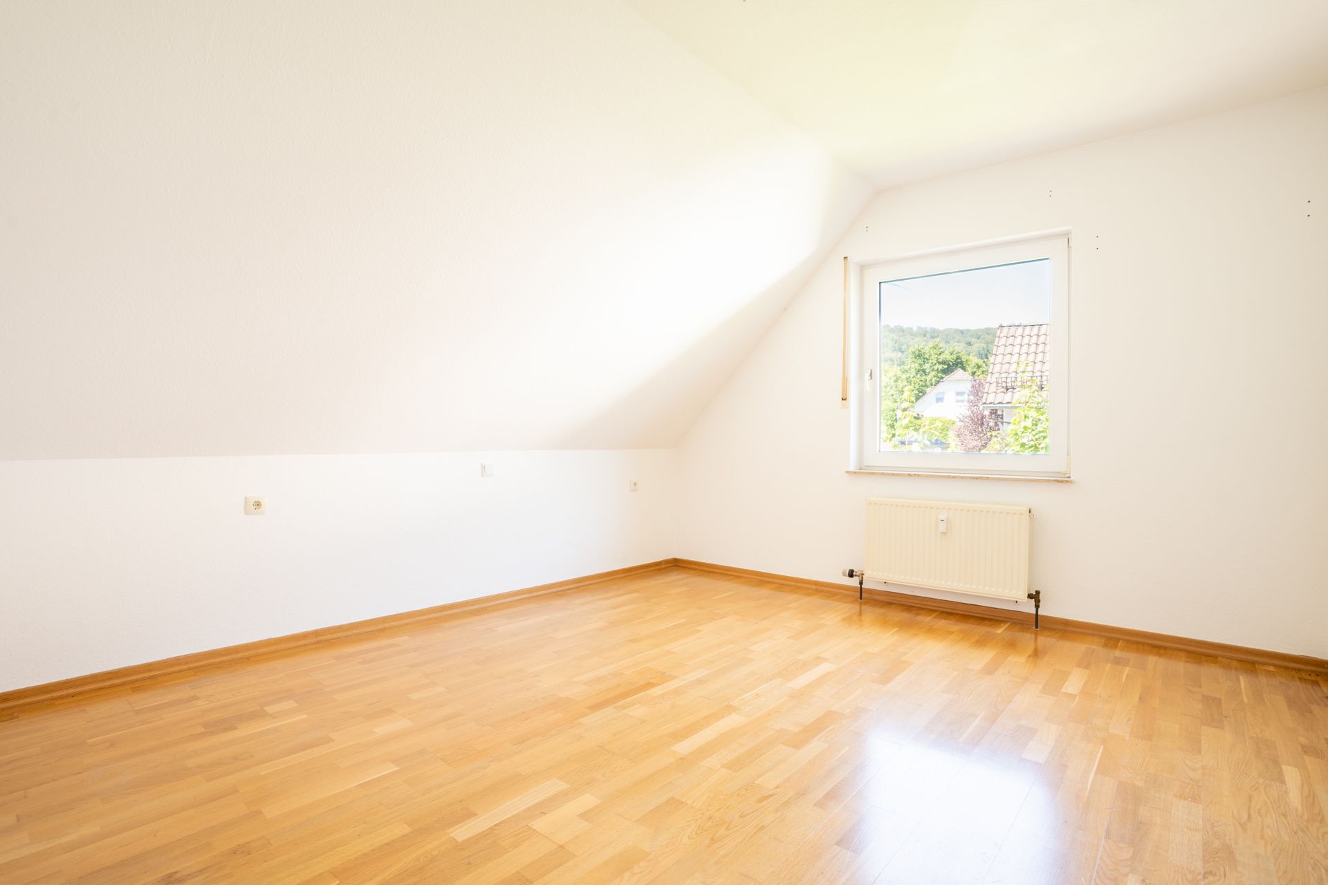 Helles, leeres Zimmer mit Holzfußboden, weißer Wand und Fenster mit Blick nach draußen.