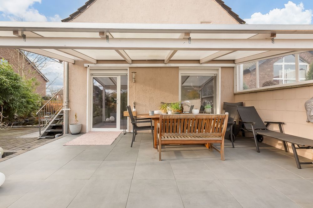 Überdachte Terrasse mit Holzmöbeln und Pflanzendekoration vor einer Wohnhaustür.