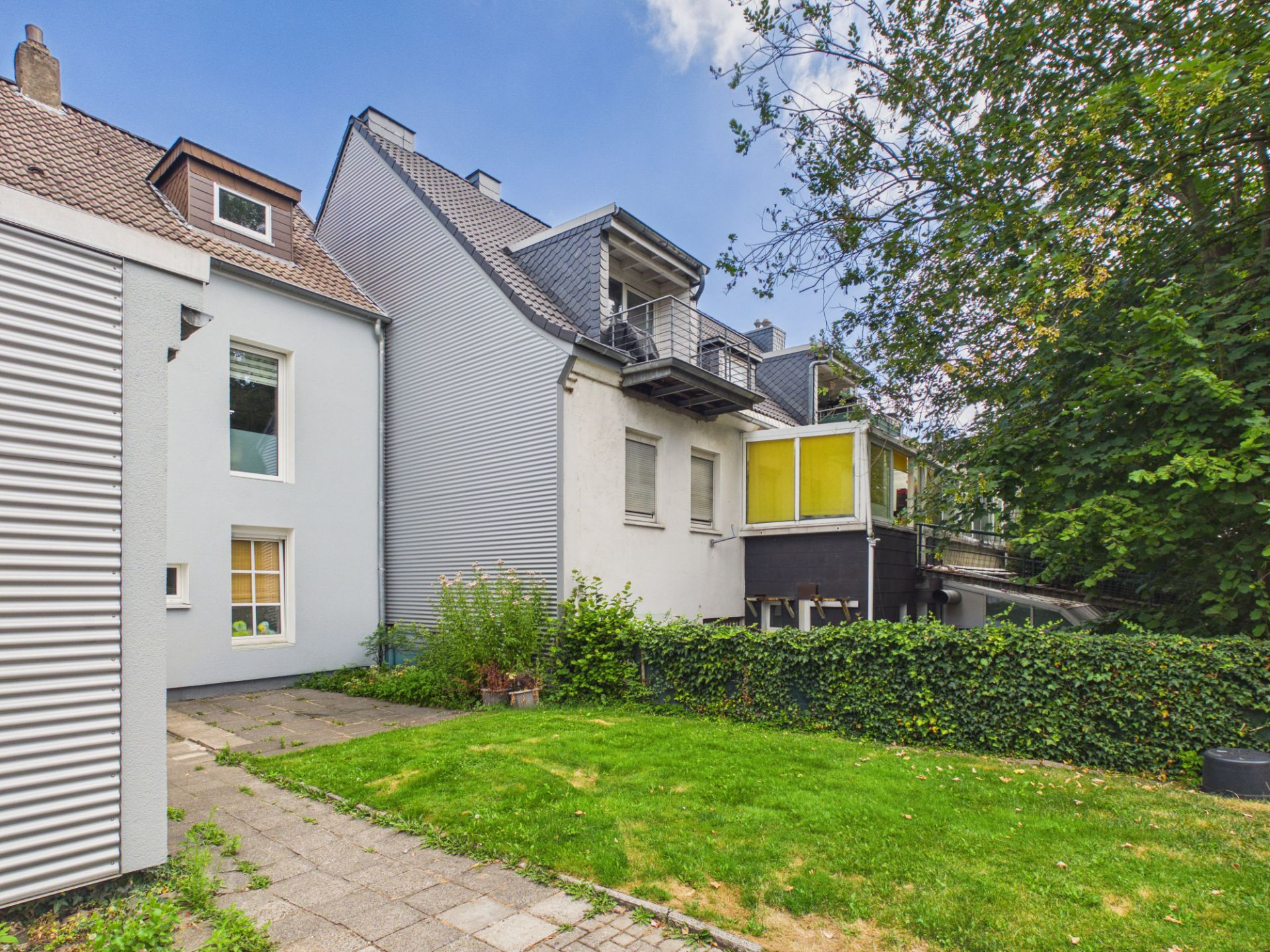 Mehrfamilienhaus mit Balkon, Garten und gepflastertem Weg, umgeben von Büschen und Bäumen.