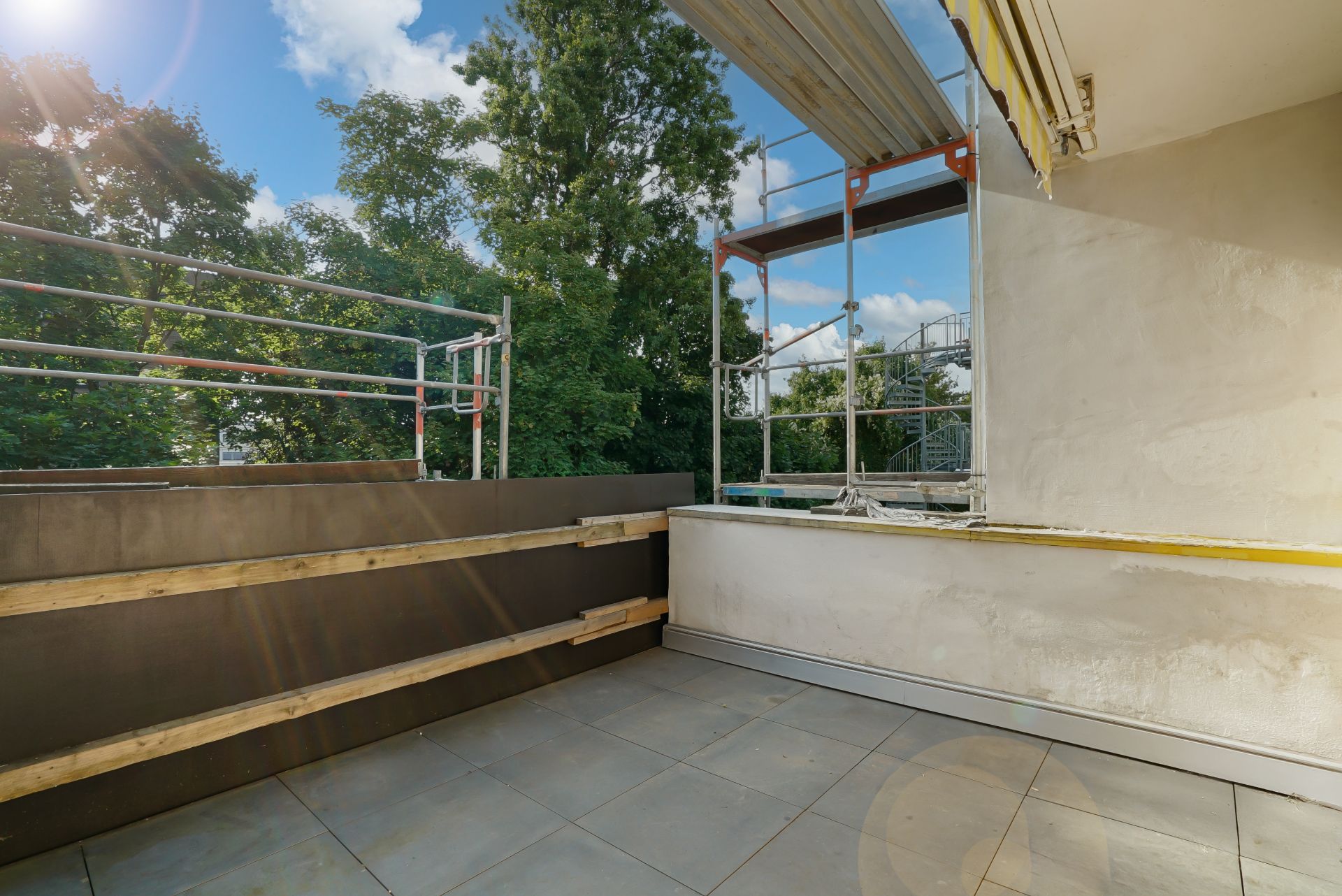 Balkon mit Fliesenboden und Baugerüst, im Hintergrund Bäume und blauer Himmel.