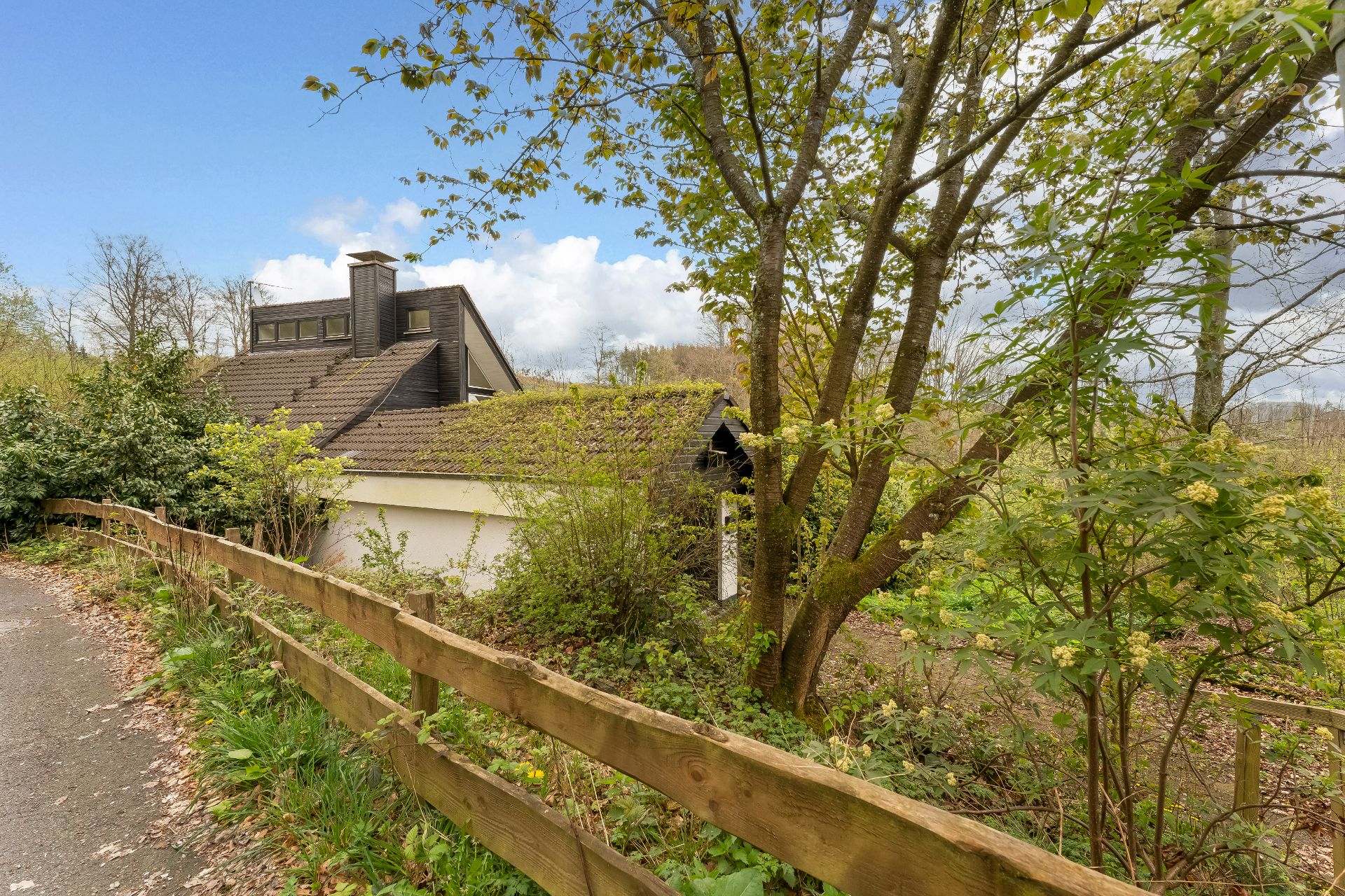 Haus mit Satteldach, von Bäumen und Sträuchern umgeben, Holzzaun im Vordergrund.