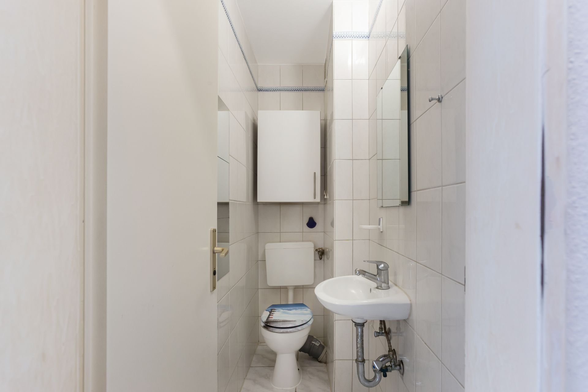Kleines Badezimmer mit Toilette, Waschbecken, Spiegel und weißen Fliesen.