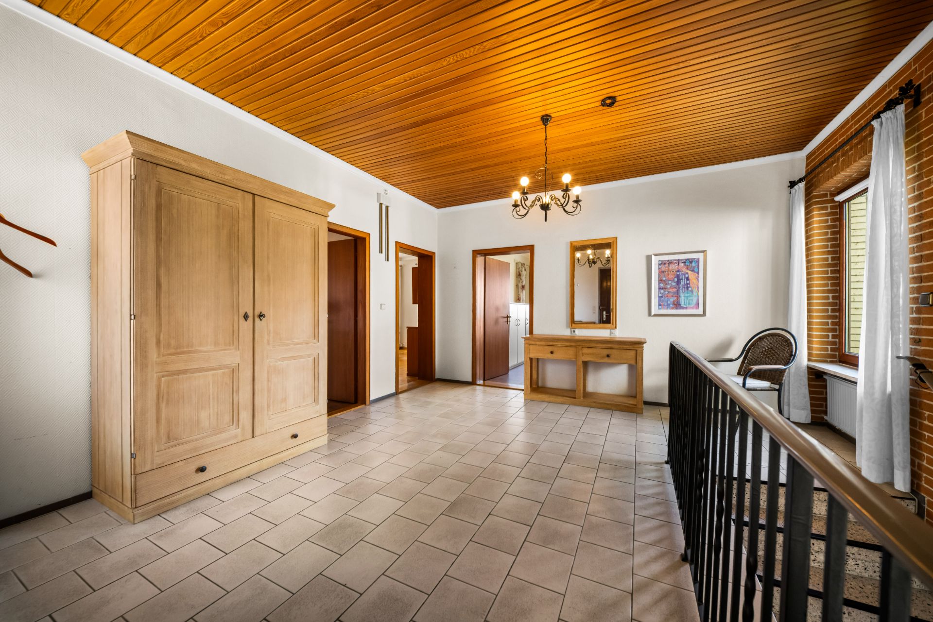 Heller Flur mit Fliesenboden, Holzdecke, Kleiderschrank und Kommode, Geländer und Fenstern.