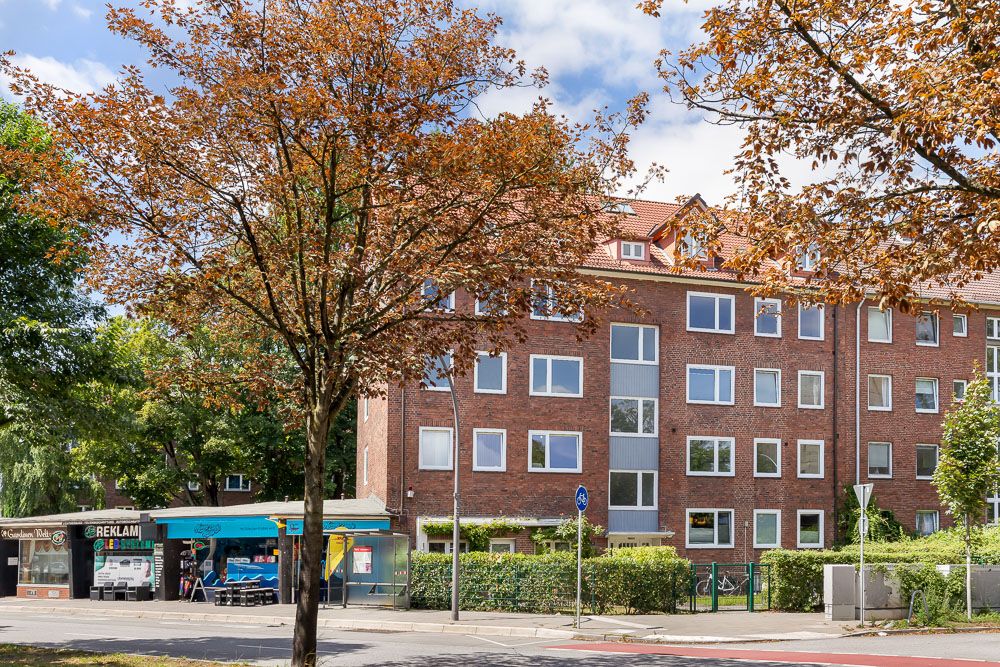 Mehrstöckiges Wohnhaus mit roten Ziegeln, Bäumen und kleinen Geschäften im Vordergrund.
