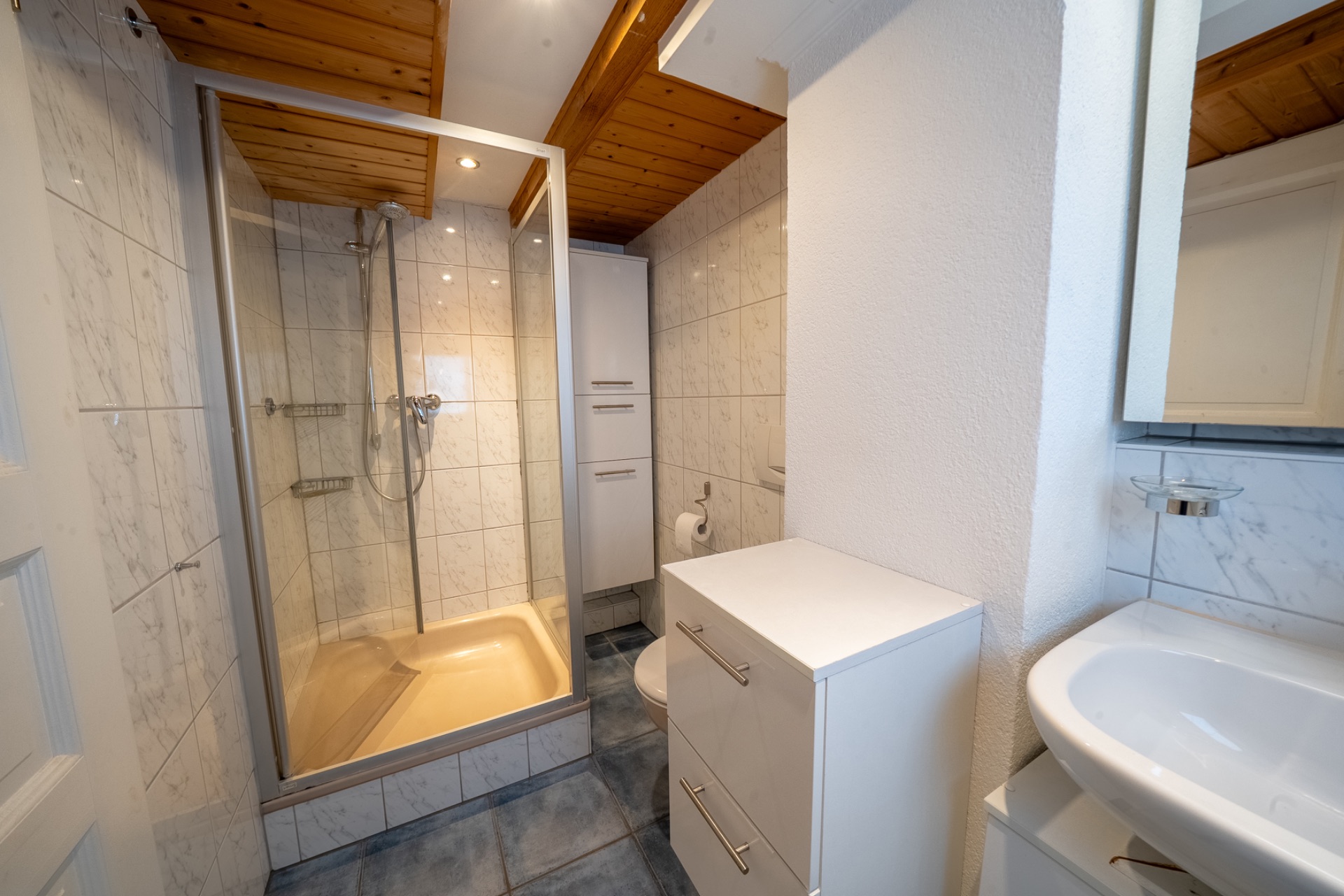 Modernes Badezimmer mit Dusche, Waschbecken, Schrank, WC und Holzdecke.