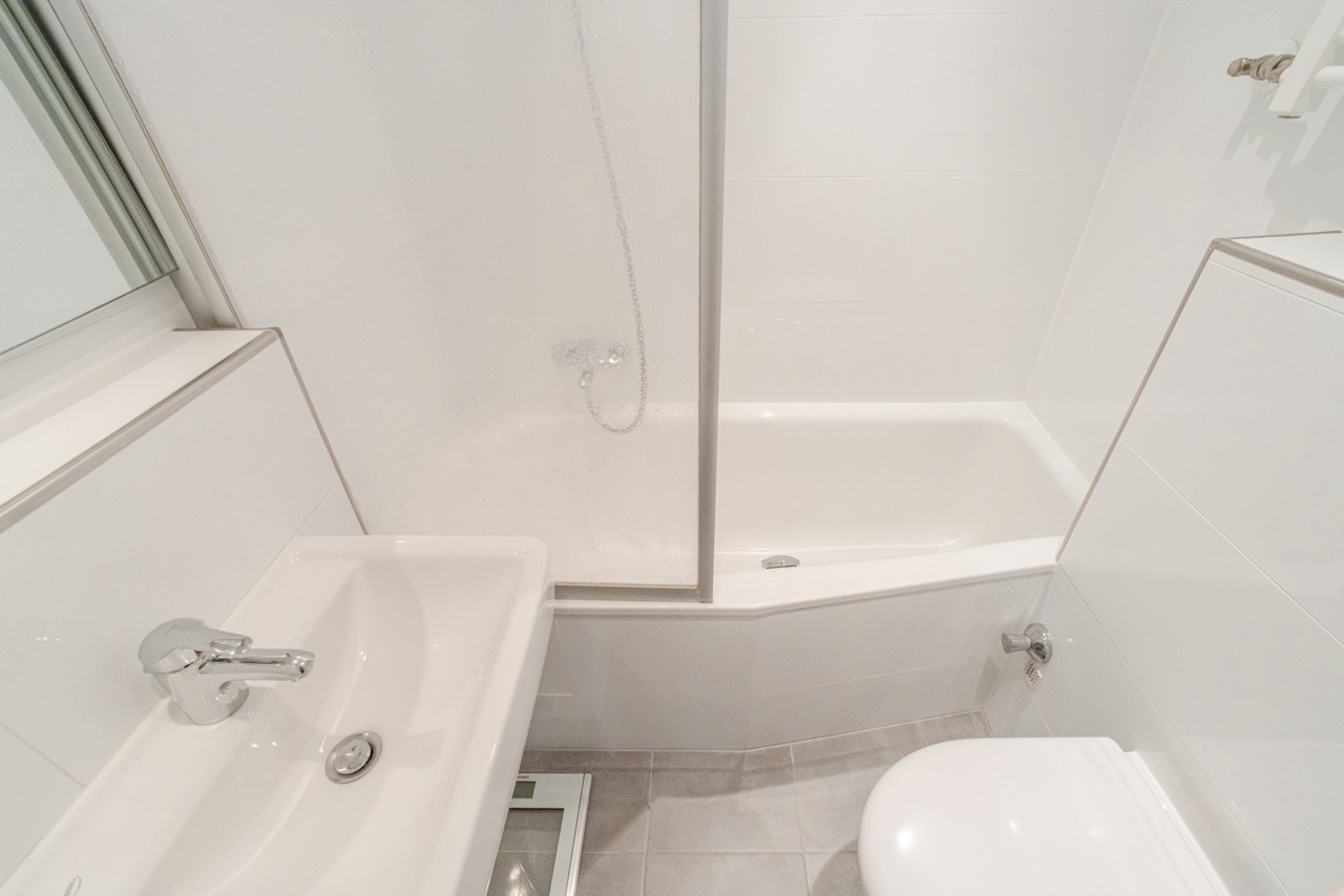 Kleines, weiß gefliestes Badezimmer mit Waschbecken, Badewanne und Toilette.