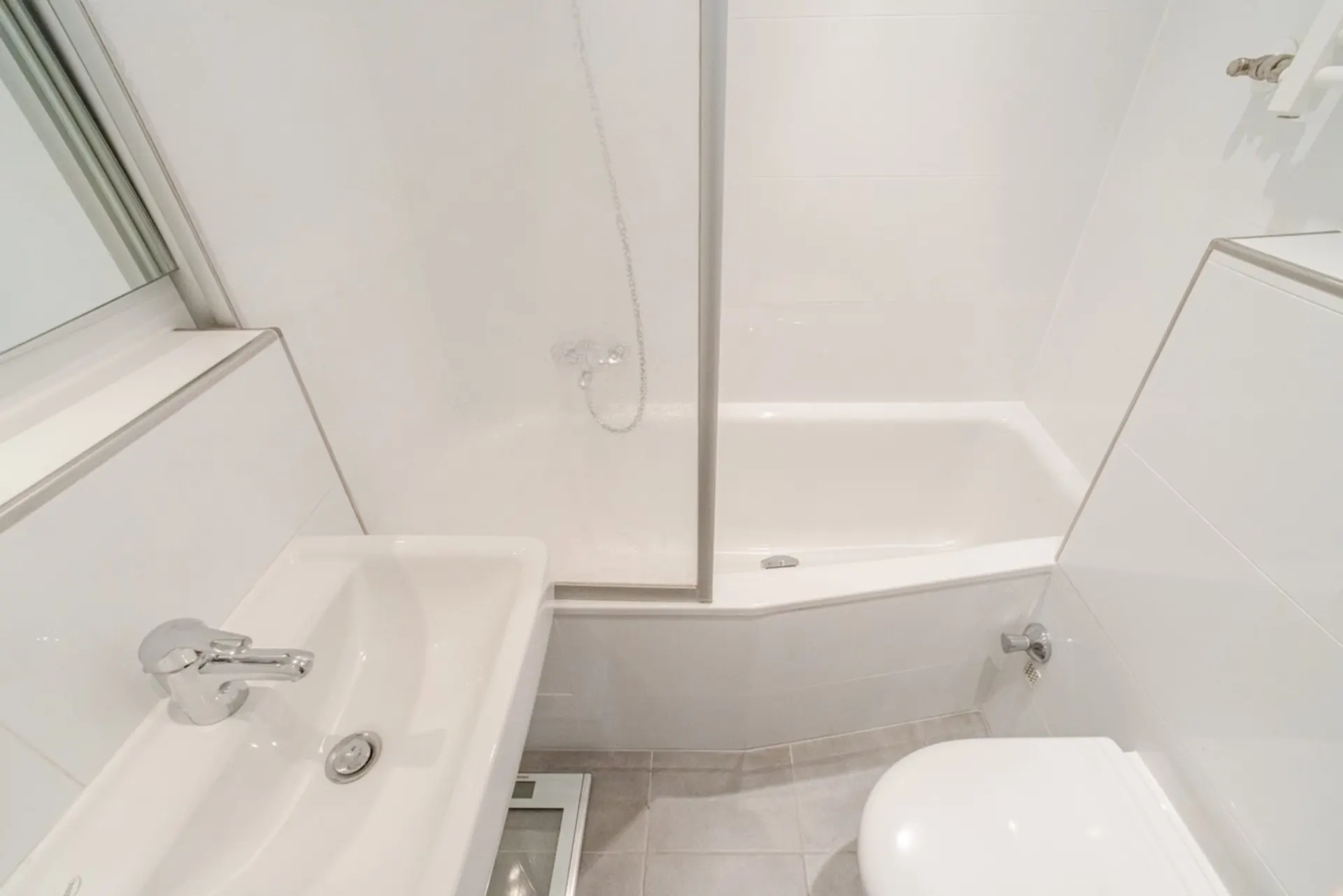 Kleines, weiß gefliestes Badezimmer mit Waschbecken, Badewanne und Toilette.