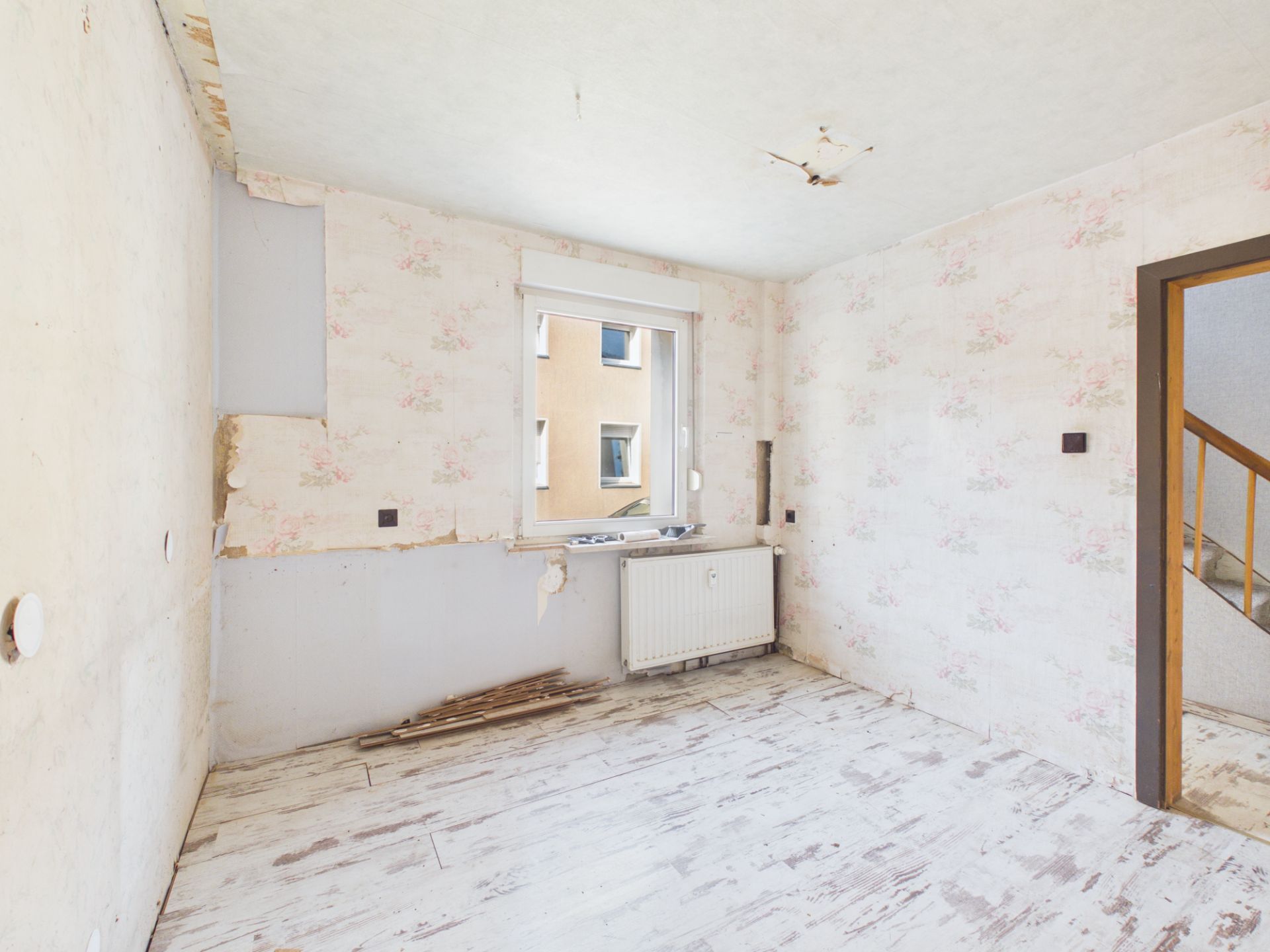 Leerer, renovierungsbedürftiger Raum mit Fenster, Heizung und altem Bodenbelag.