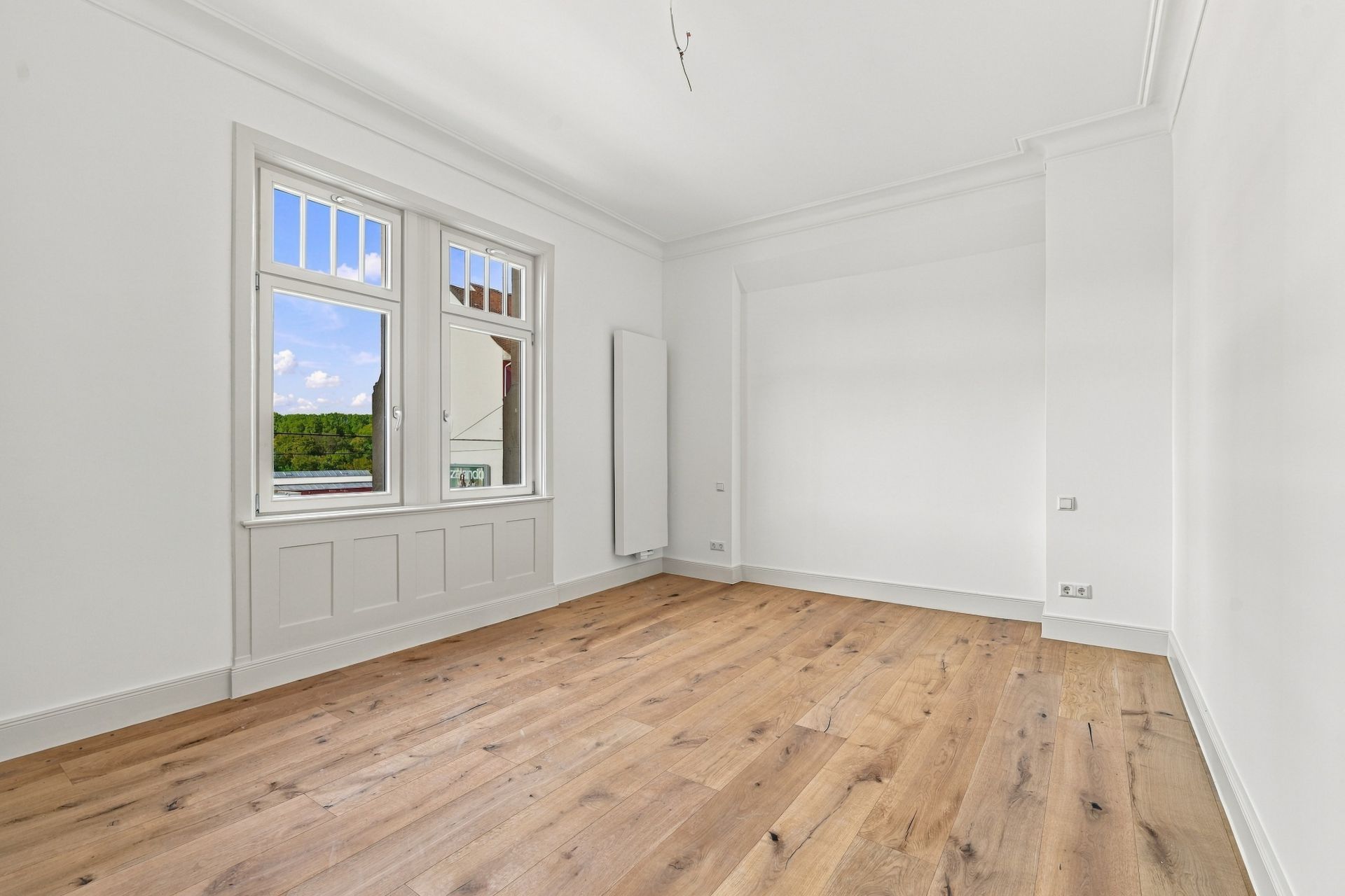 Leerer Raum mit Holzboden, weißen Wänden und einem Fenster mit Blick ins Grüne.