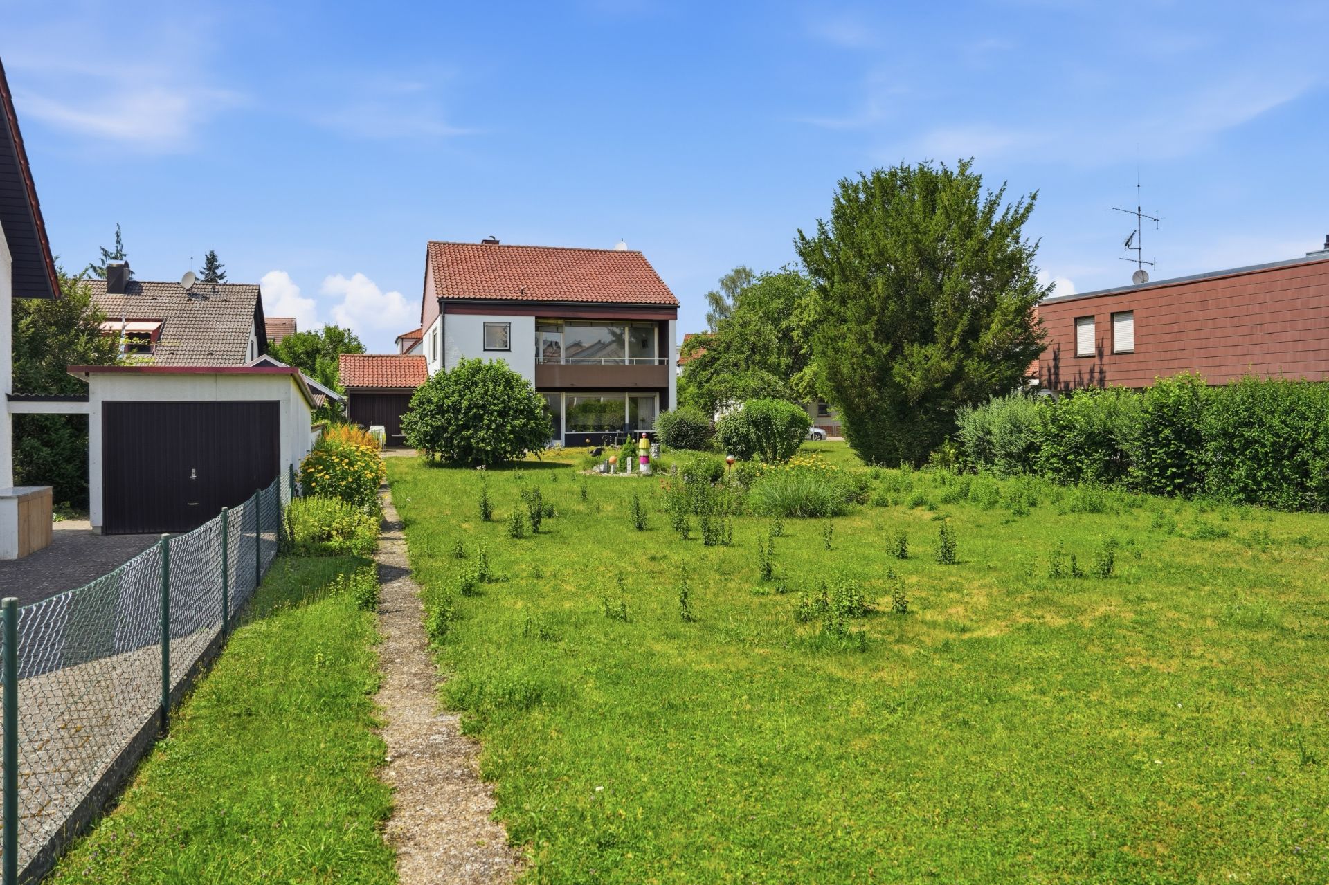 Großer grüner Garten mit Weg, links Garage, im Hintergrund ein Wohnhaus mit Balkon.
